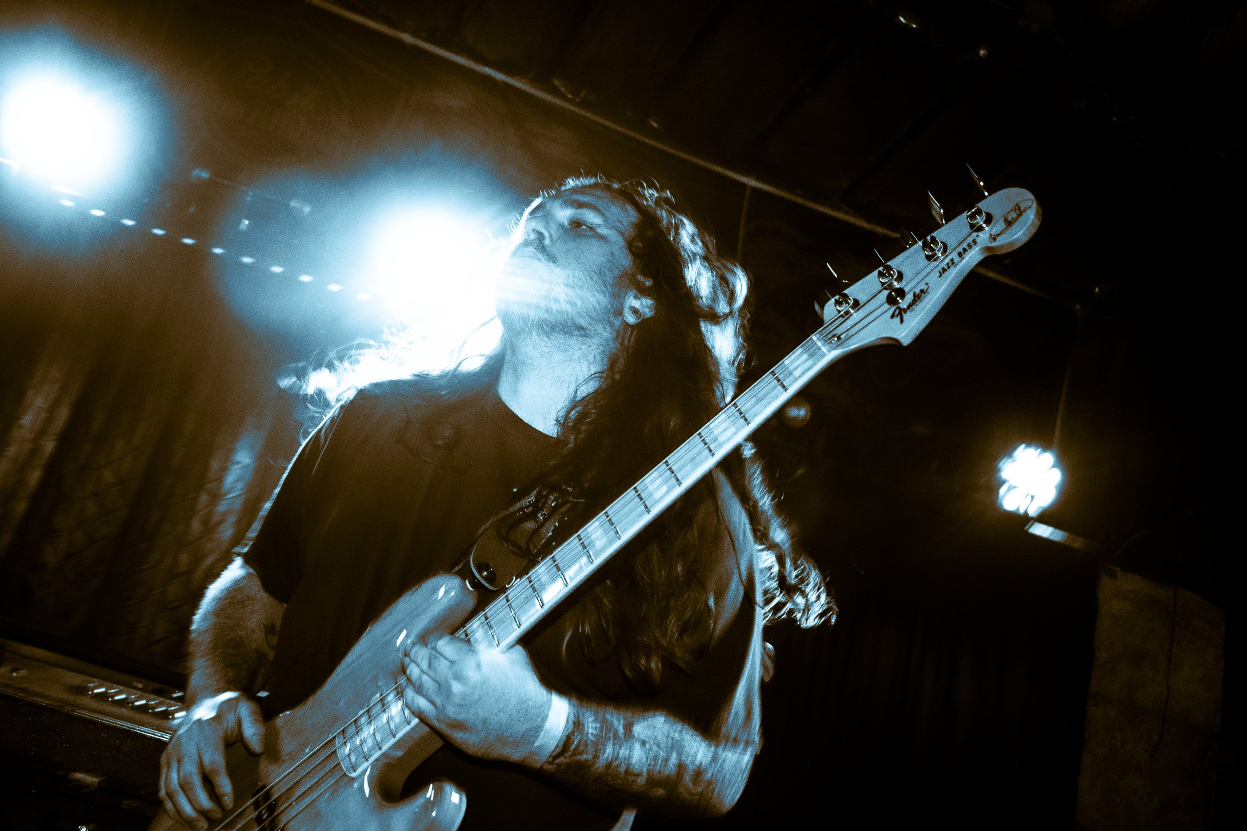 Tysonious Mink of the band Liquid Pennies playing a Fender Jazz Bass guitar on stage, illuminated by blue stage lights.