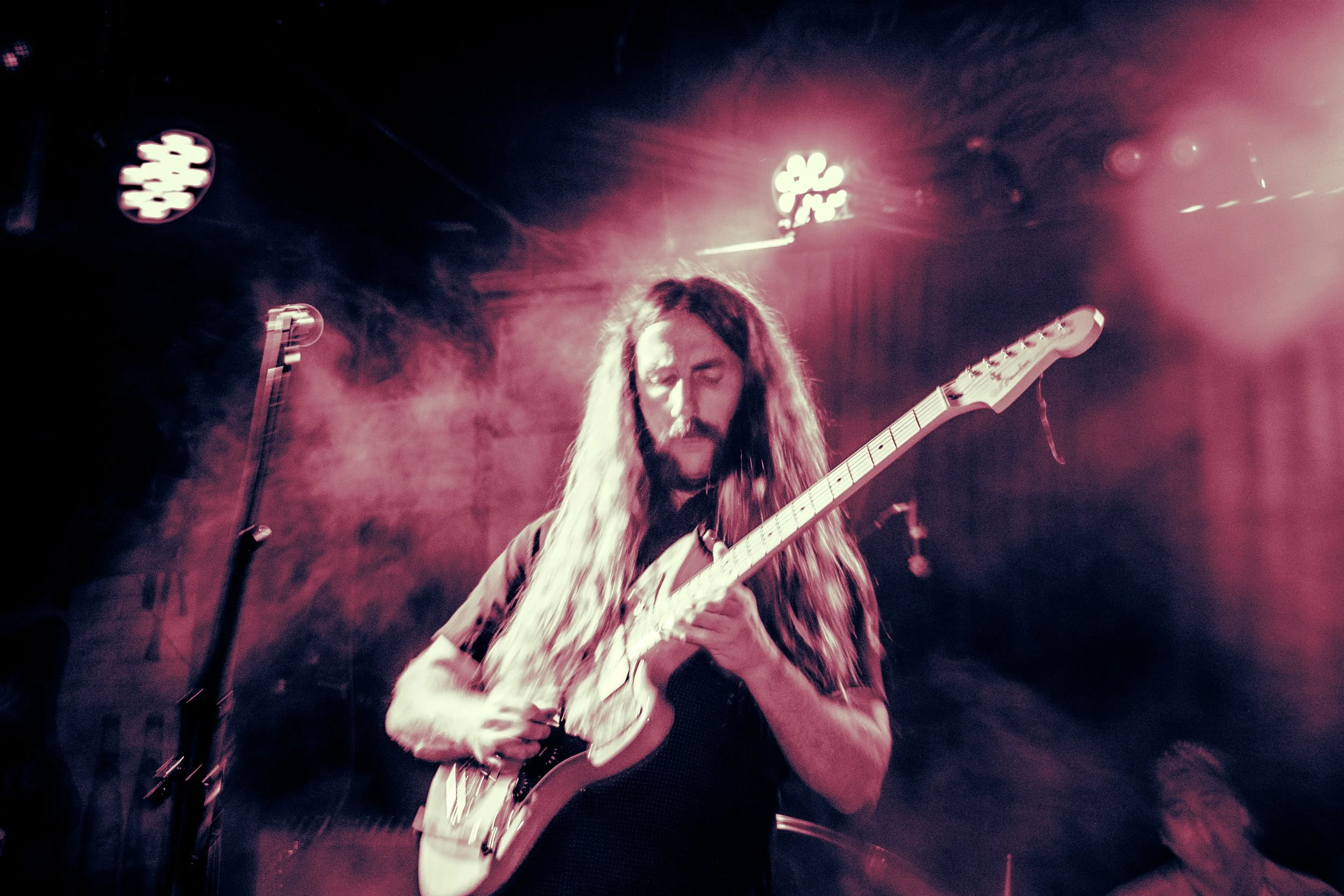 Rock band Liquid Pennies frontman/guitarist Chas BInns, a male guitarist with long hair playing electric guitar on stage with pink lighting and smoke effects.
