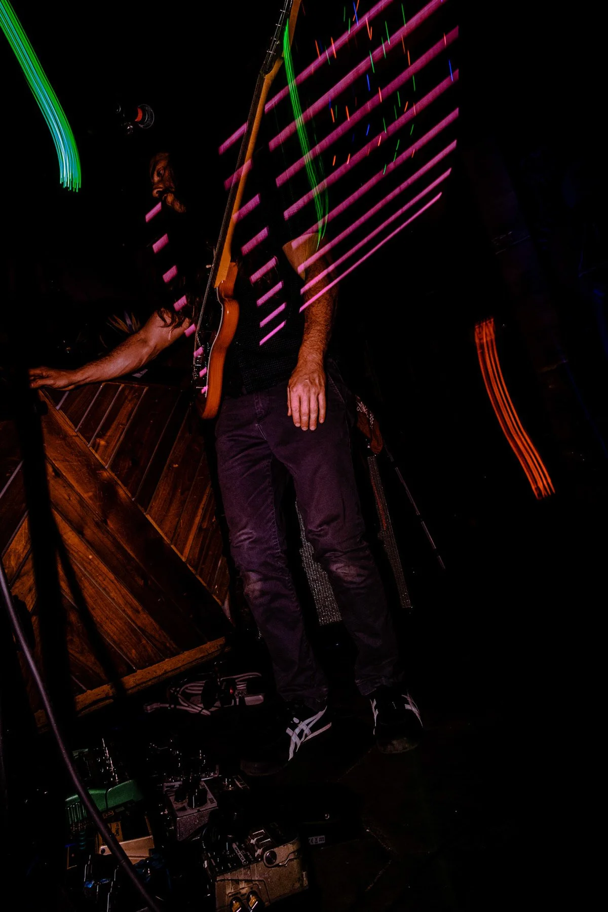 A musician playing a guitar on stage, surrounded by colorful light trails and stage equipment, with a dark background.