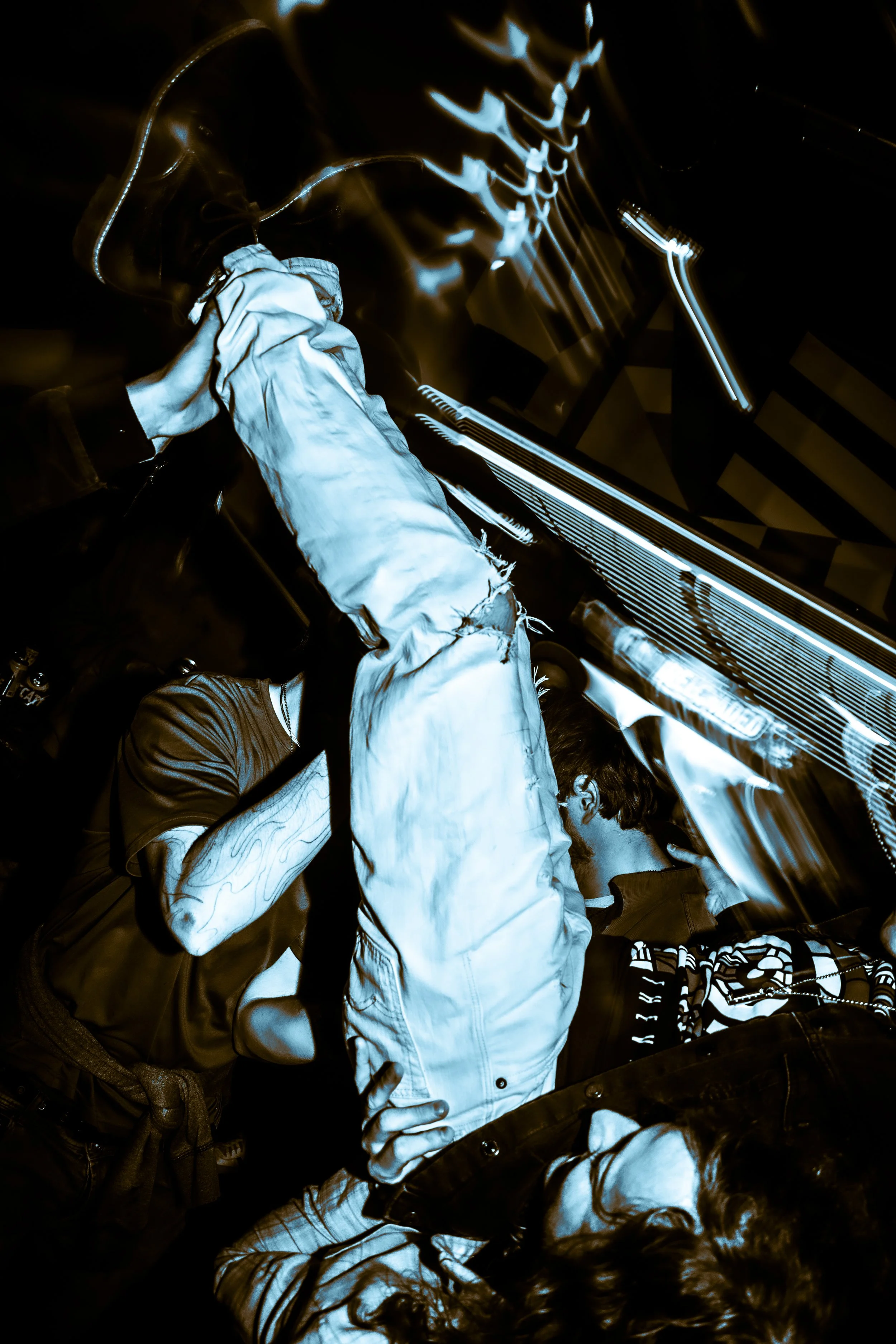 Two people are in a nightclub with blue lighting, one wearing a distressed white jacket and the other with patterned clothing, standing near a bar or booth, with motion blur and light streaks creating a dynamic, energetic atmosphere.