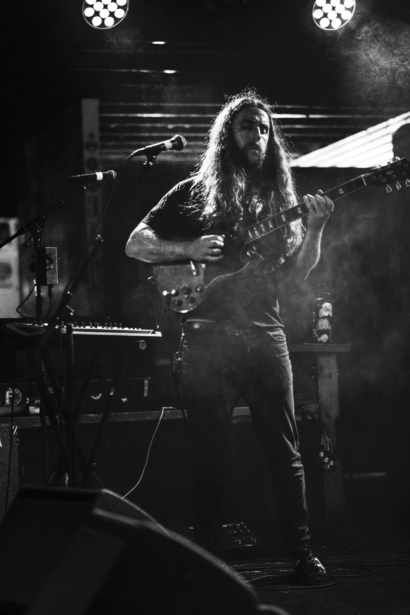 A musician with long hair and a beard playing electric guitar on stage with microphones and musical equipment around him, illuminated by stage lights.