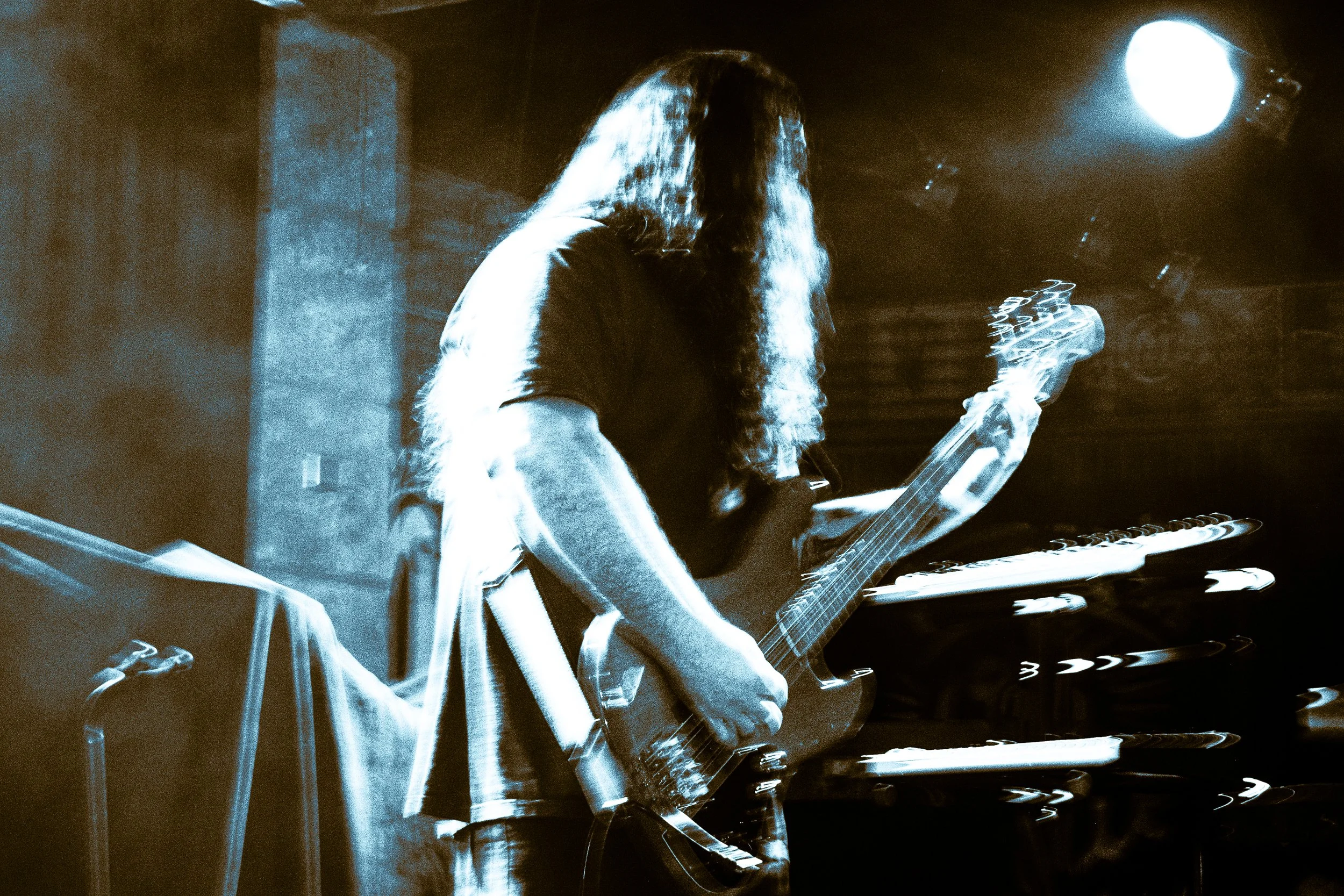A musician with long hair playing an electric guitar on stage, with stage lights creating streaks of light and a blurred background.