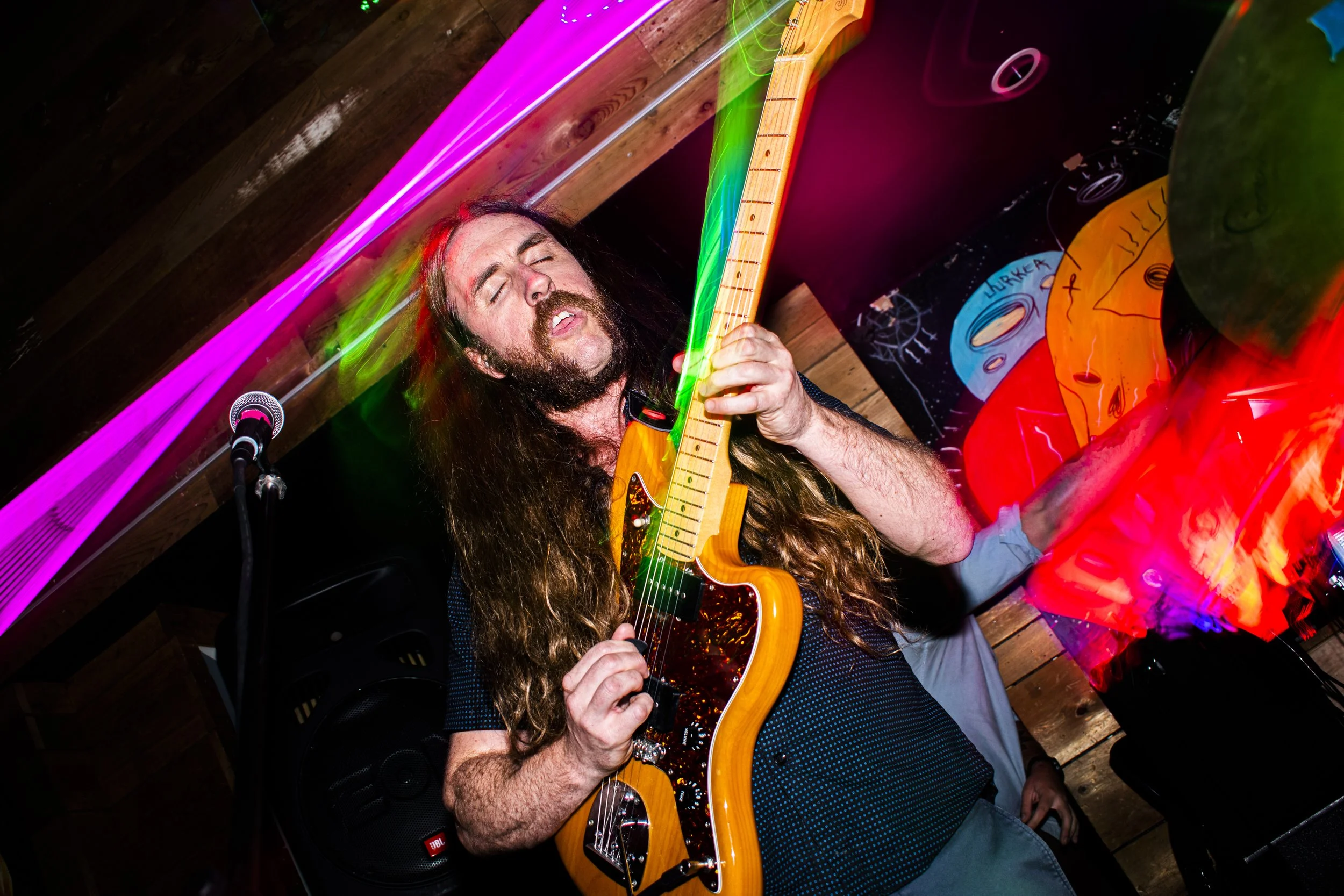 Liquid Pennies frontman/guitarist Chas Binns playing an electric guitar with eyes closed. The setting includes colorful neon lights and wooden wall panels, suggesting a lively performance at a music venue.