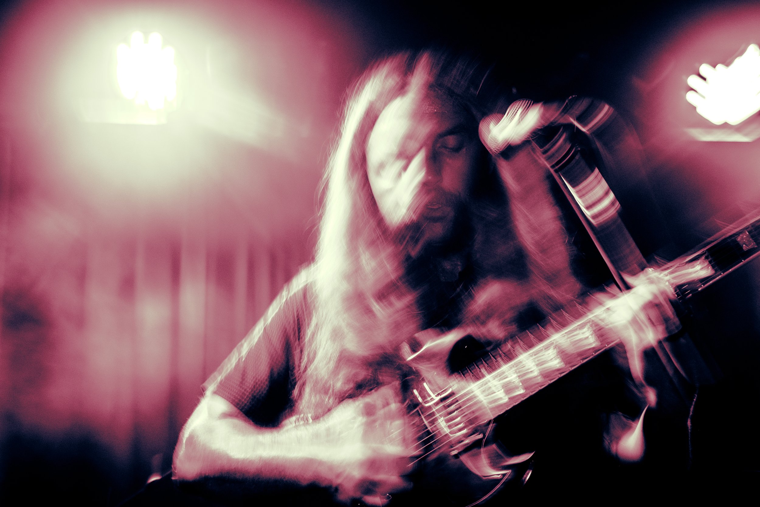 Rock band Liquid Pennies frontman/guitarist Chas BInns, with long hair, playing an electric guitar, with motion blur and pink lighting, creating an abstract, energetic scene.
