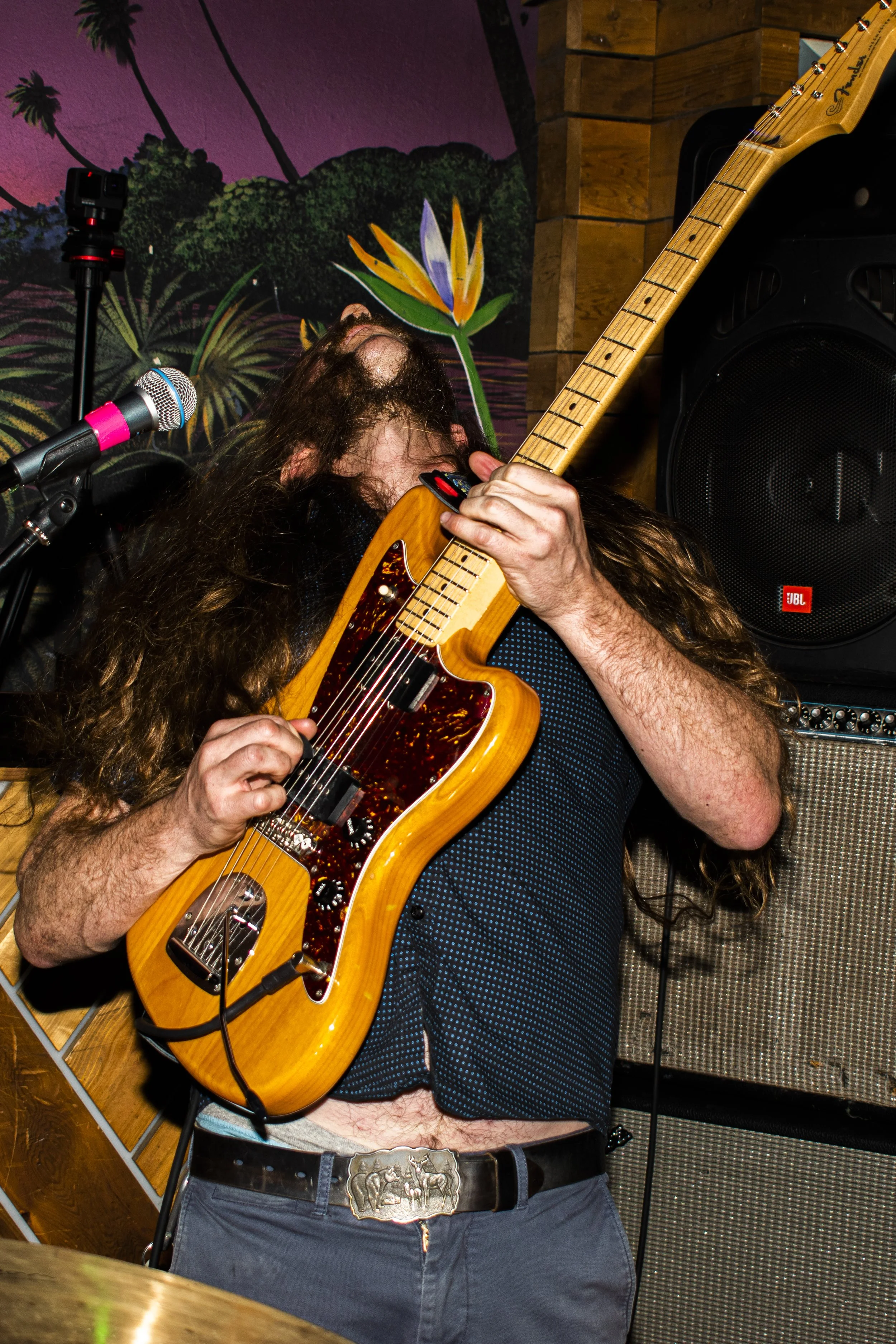 Musician with long hair playing a yellow electric guitar with a tortoise shell pickguard on a stage decorated with a mural of tropical plants and a sunset background.