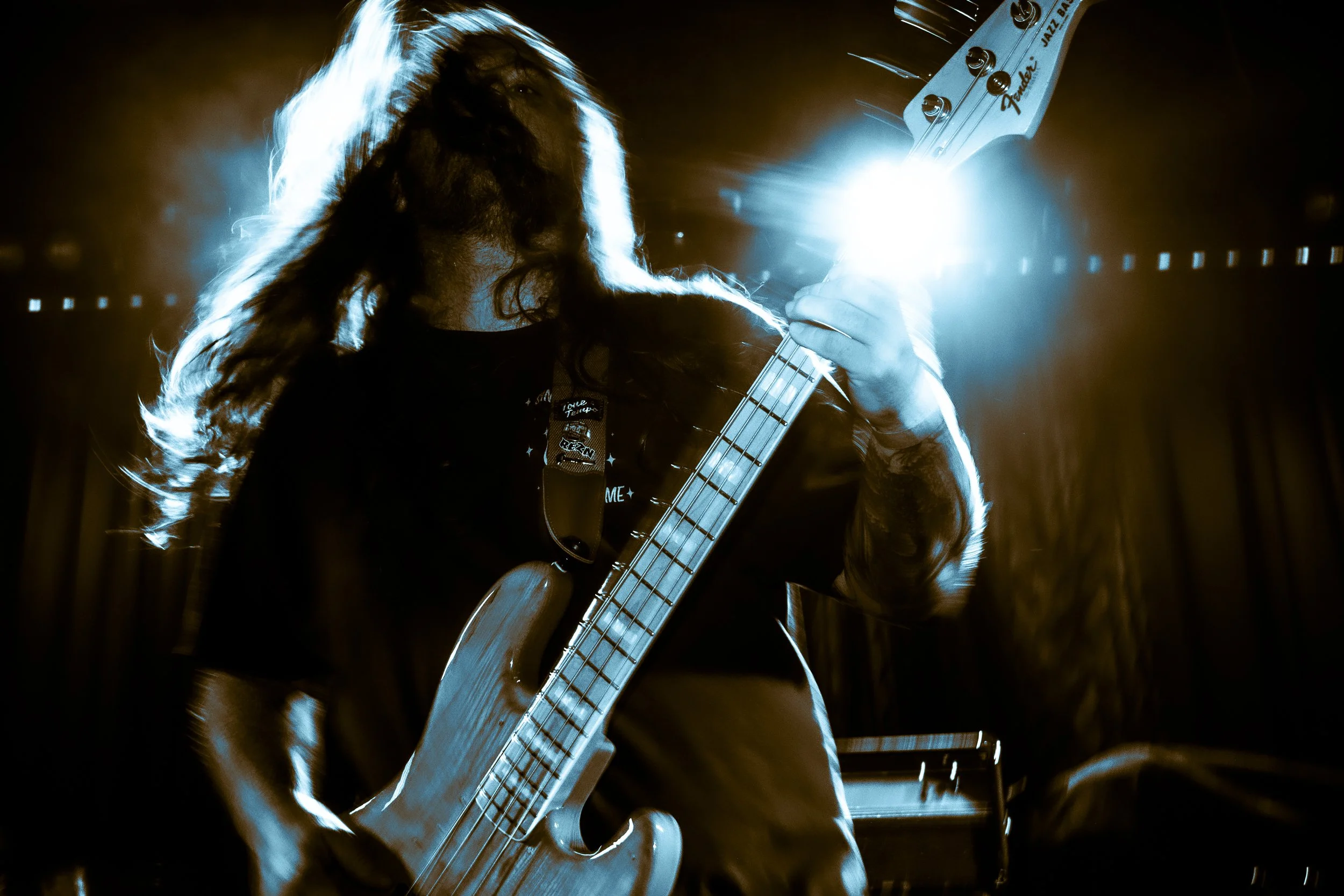 A musician with long hair playing a guitar on stage, backlit by bright blue and orange lights, creating a silhouette effect.