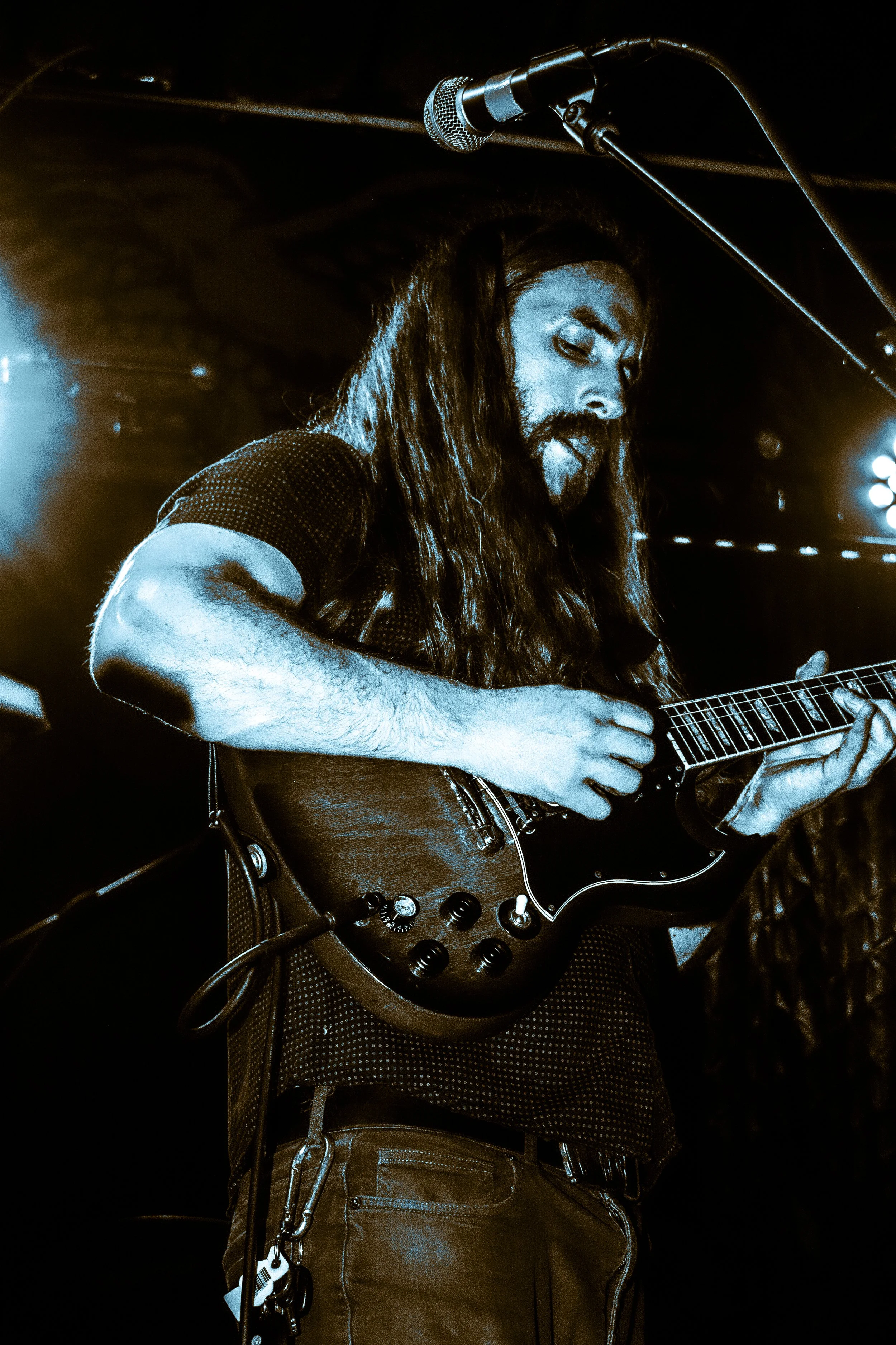 A male musician with long hair and beard playing an electric guitar on stage near a microphone during a performance.