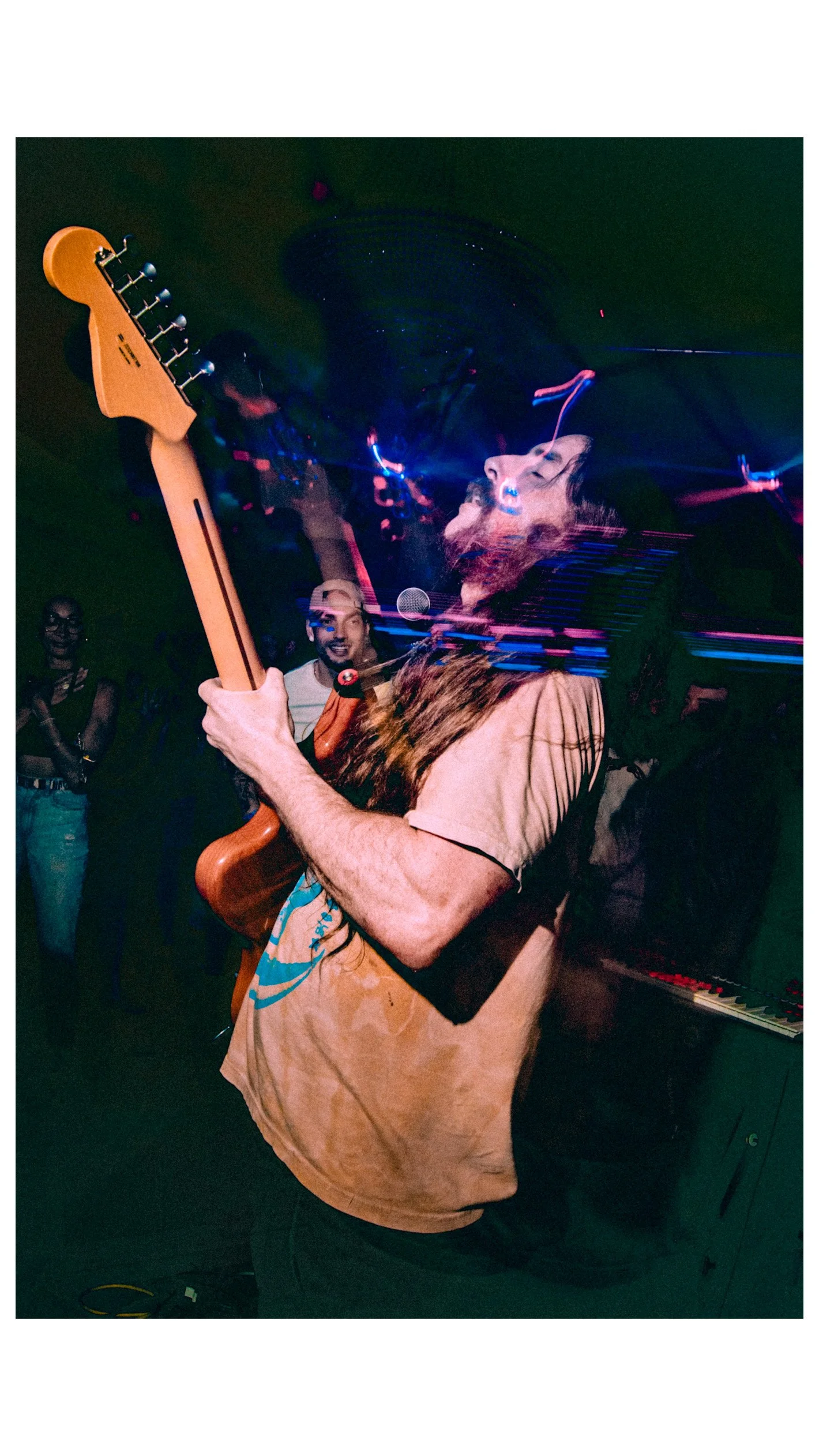 Chas Binns of the band Liquid Pennies playing an electric guitar on stage, with a colorful light display in the background and a woman with glasses in the audience.