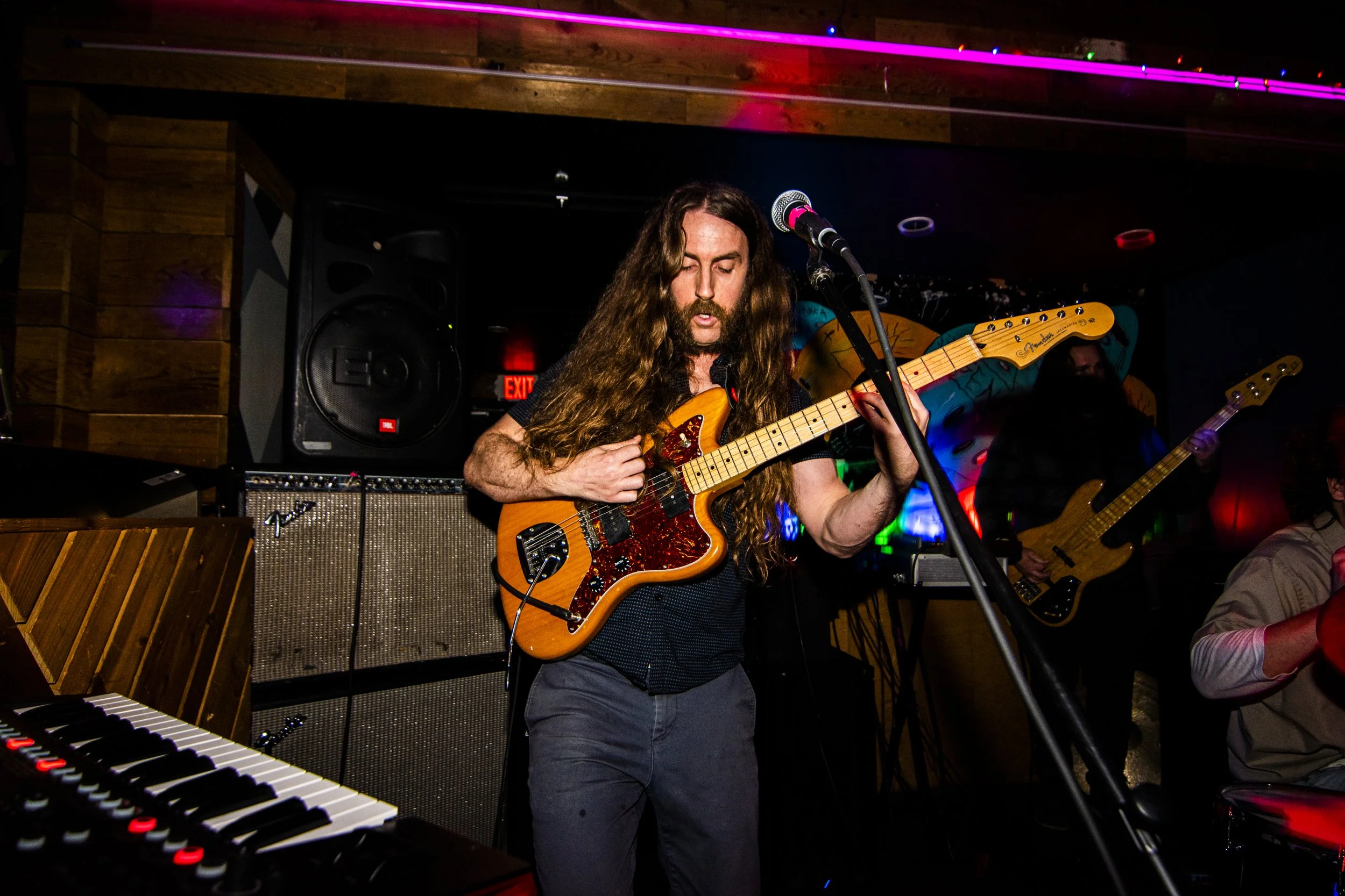 A man with long, curly hair playing an orange electric guitar in a dimly lit music venue. There are musical equipment, a keyboard, and other band members in the background.