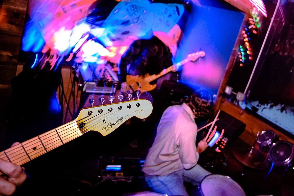 Two musicians playing electric guitars and a drummer in a colorful, dimly lit room with neon lights and decorated walls.