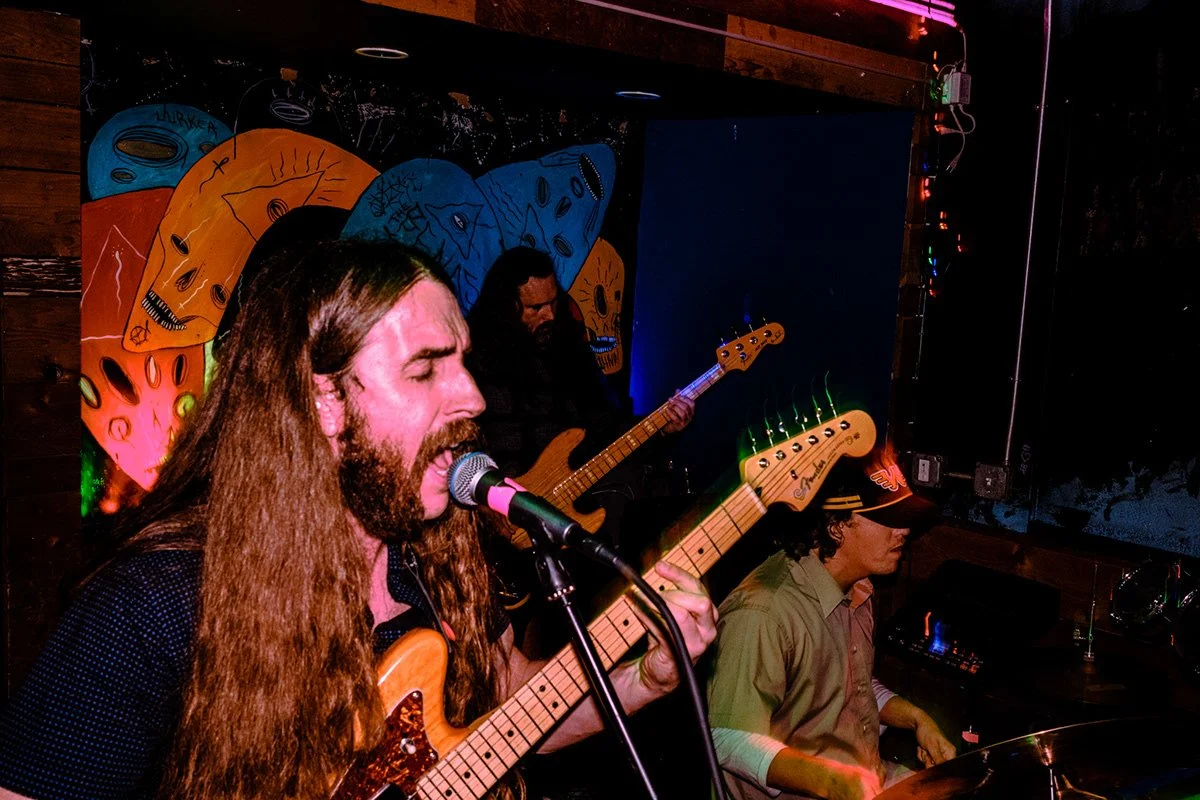 A band performing live music at a venue with colorful artwork of faces in the background; a man with long hair and a beard plays an electric guitar and sings into a microphone, a woman in a hat plays bass guitar, and another person is on keyboard.