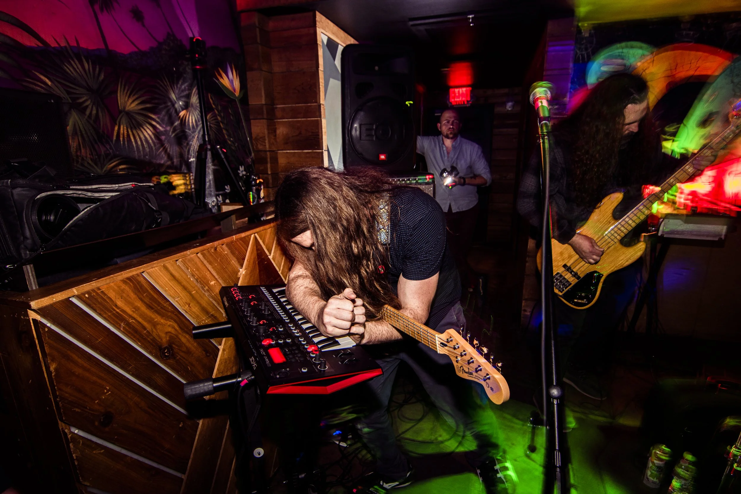 Musicians performing at a dimly lit venue, with one playing a guitar and keyboard, another playing a bass guitar, and a third in the background near DJ equipment.