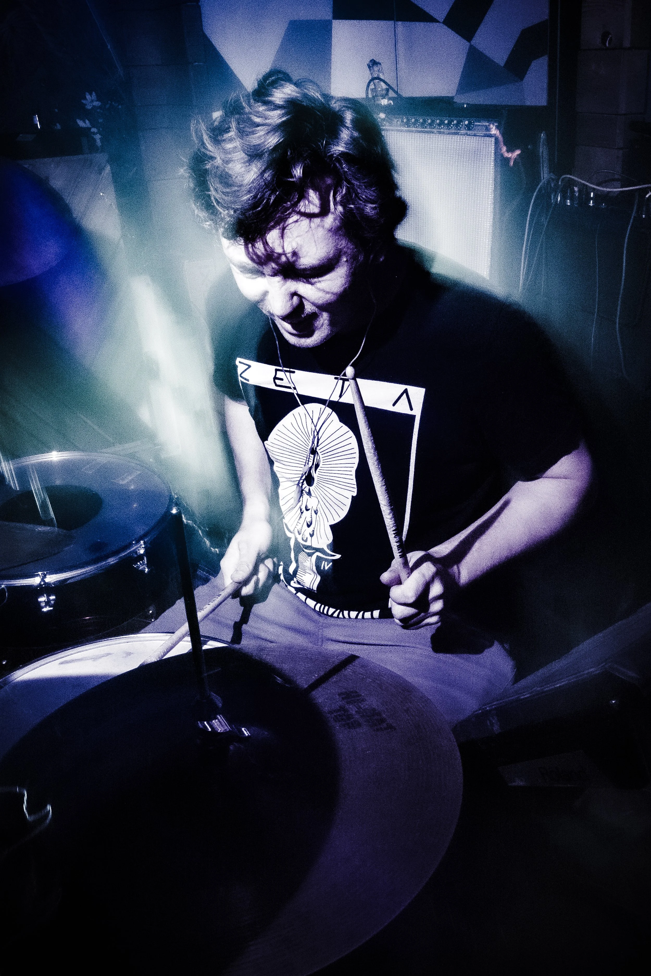 A person with curly hair intensely playing drums on a stage with colorful lighting.