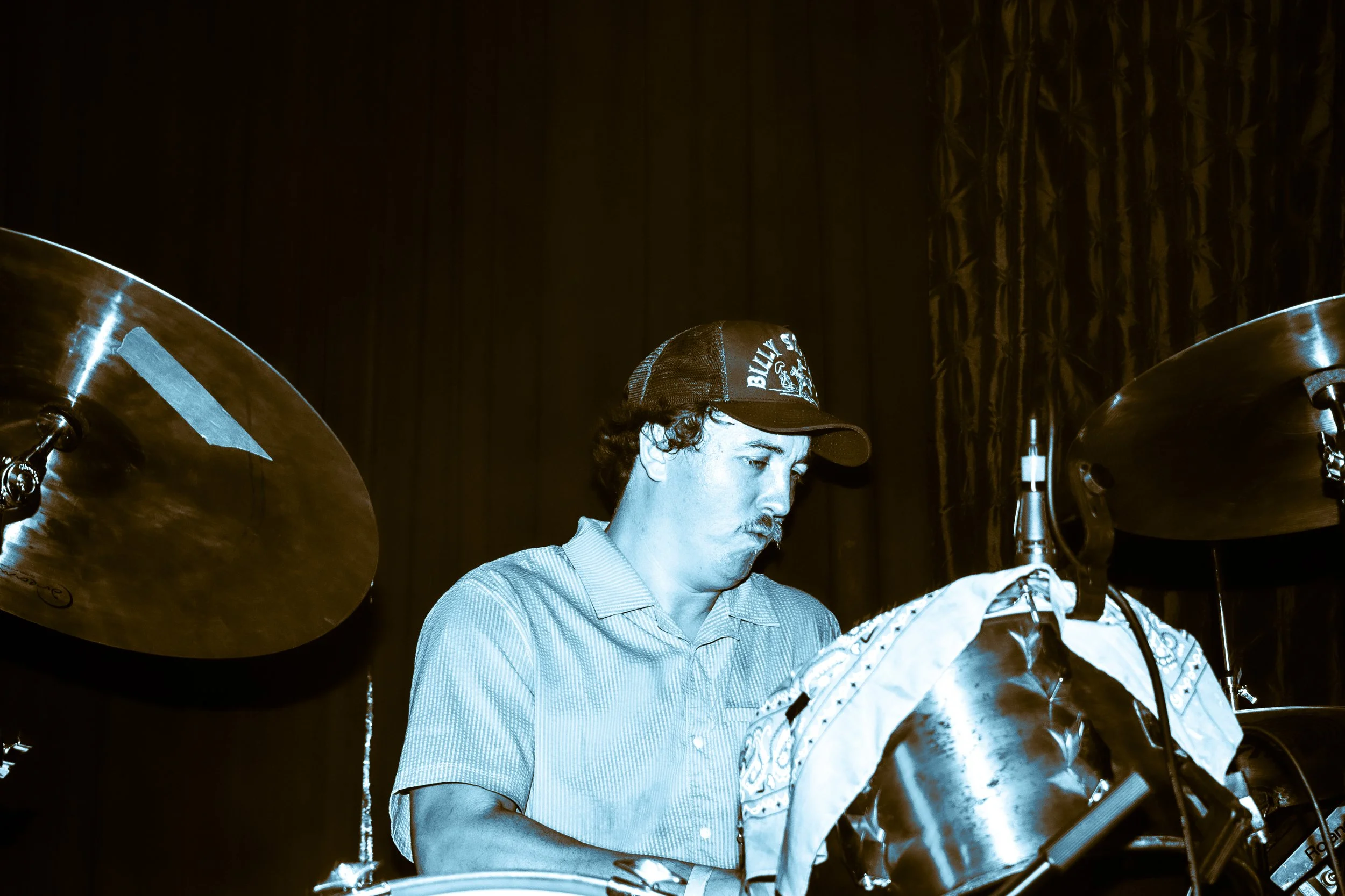 Psych rock band Liquid Pennies drummer PIerson Whicker  playing drums on stage, wearing a baseball cap, with a dark background