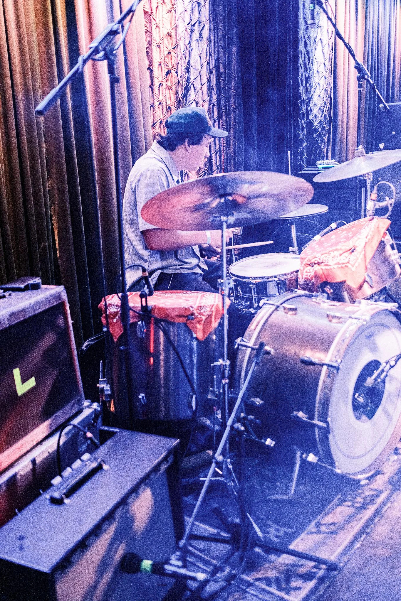 A musician playing drums on stage with a brown curtain backdrop, microphones overhead, and stage equipment nearby.