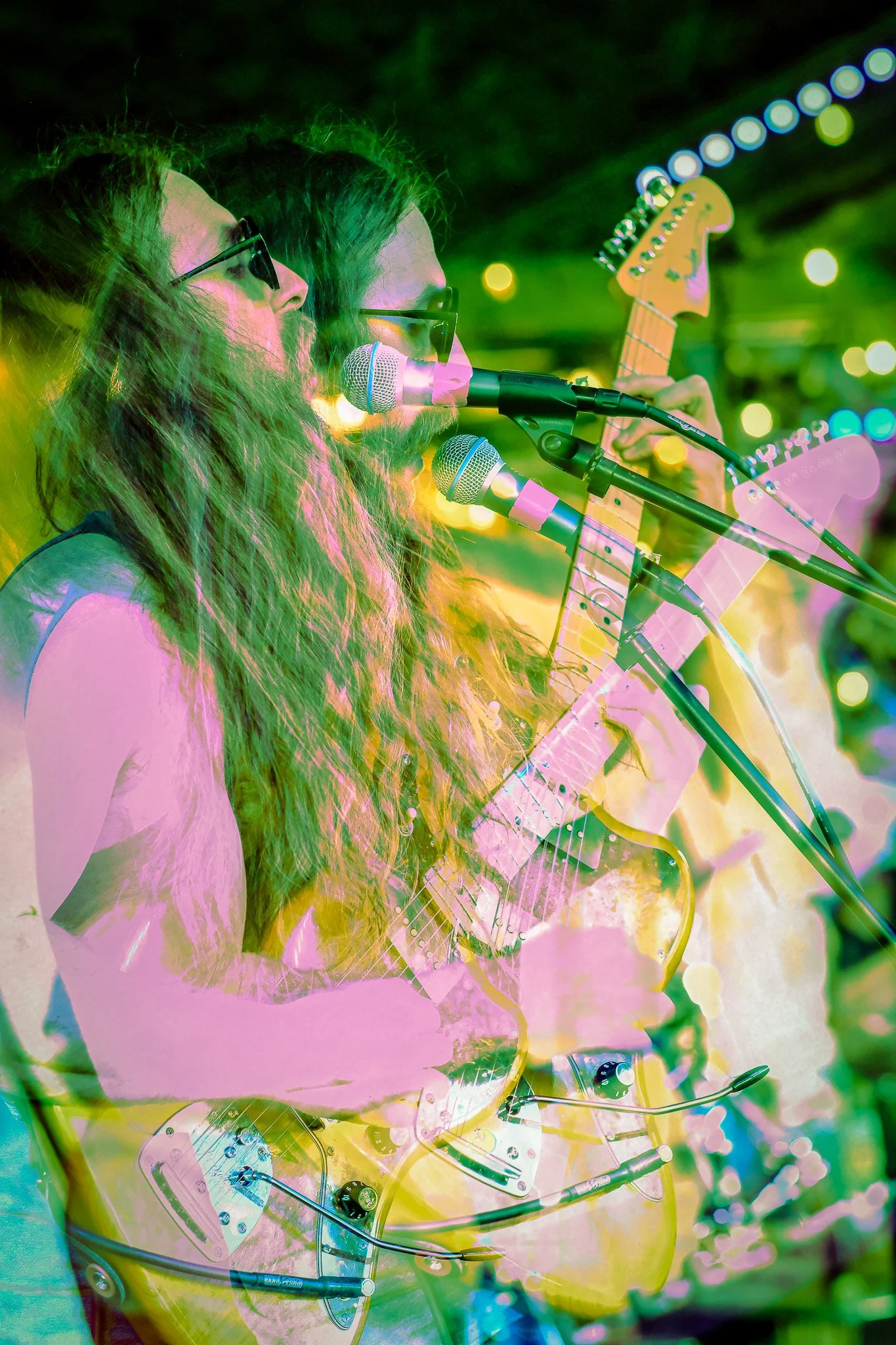 Two musicians with long hair and glasses perform with electric guitars and microphones at night under colorful lights.