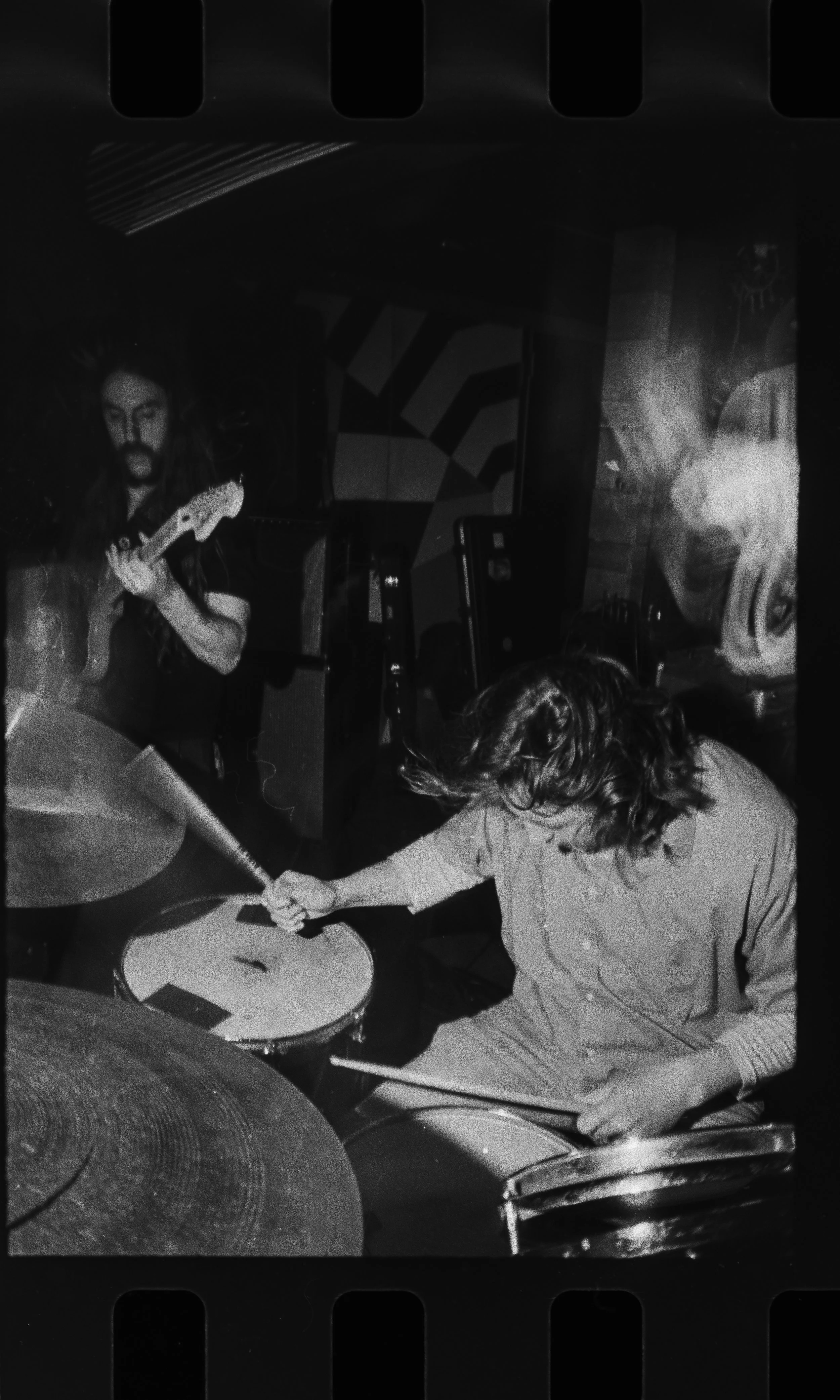 Two musicians playing instruments in a dimly lit room; one is on drums, and the other is playing guitar.