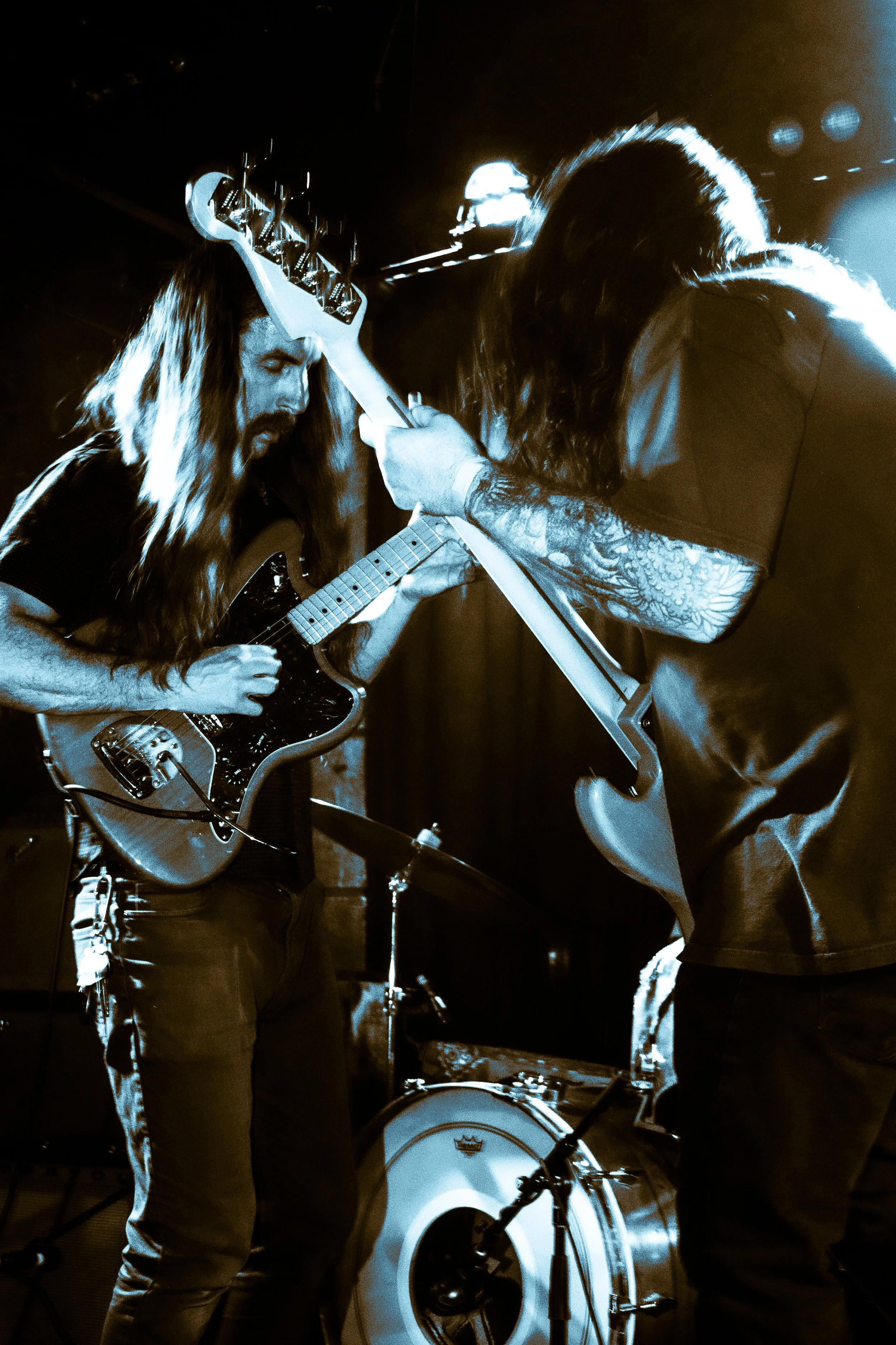 Two musicians playing electric guitars on stage, one with long hair and a beard, and the other with tattoos on their arm, under concert lighting.