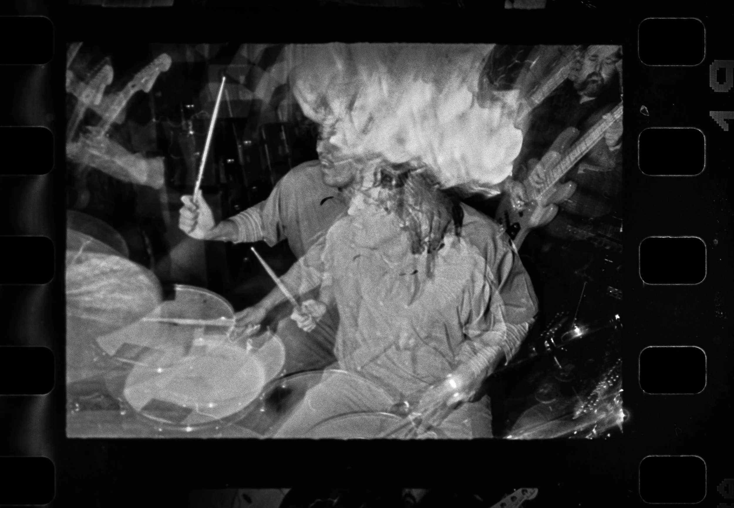 Black and white photo of a woman playing drums with a man playing guitar in the background.