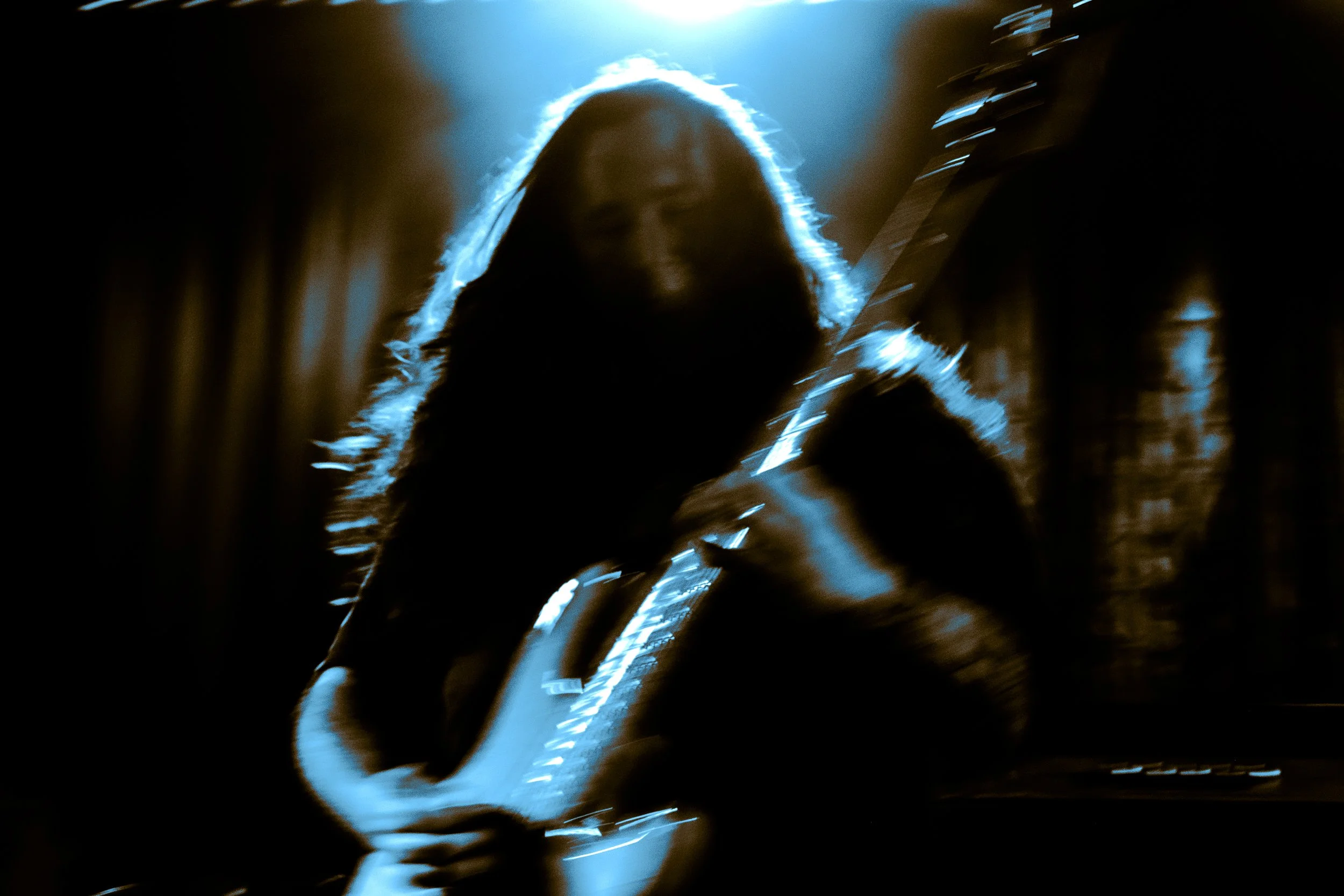 A silhouetted person with long hair playing an electric guitar, illuminated by blue light with a dark, blurred background.