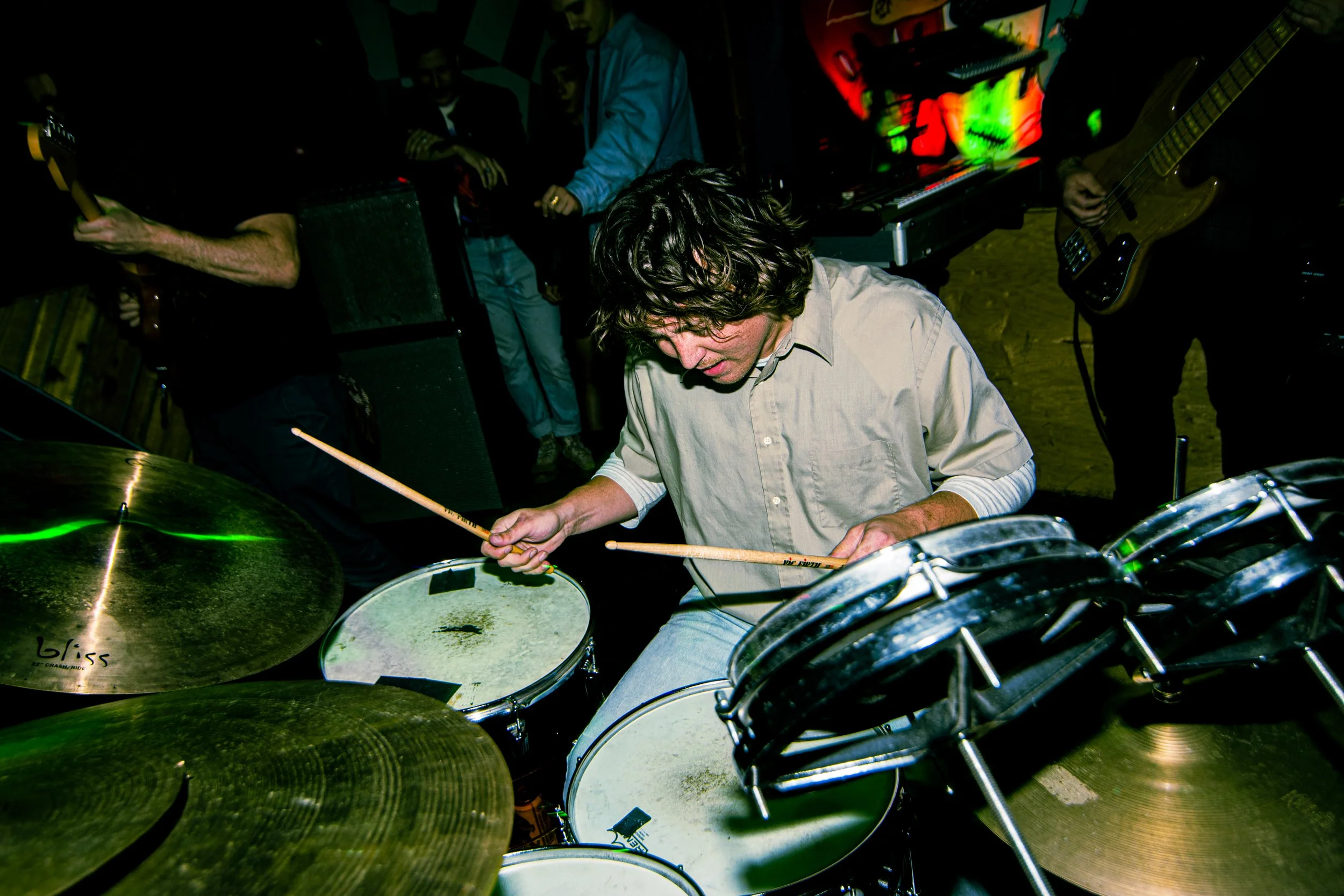 A man playing drums, surrounded by other band members and musical equipment in a dimly lit setting.