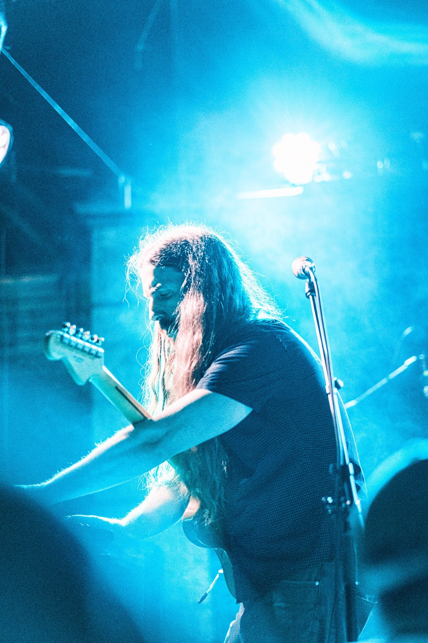 A musician with long hair playing an electric guitar on stage, illuminated by blue stage lights.
