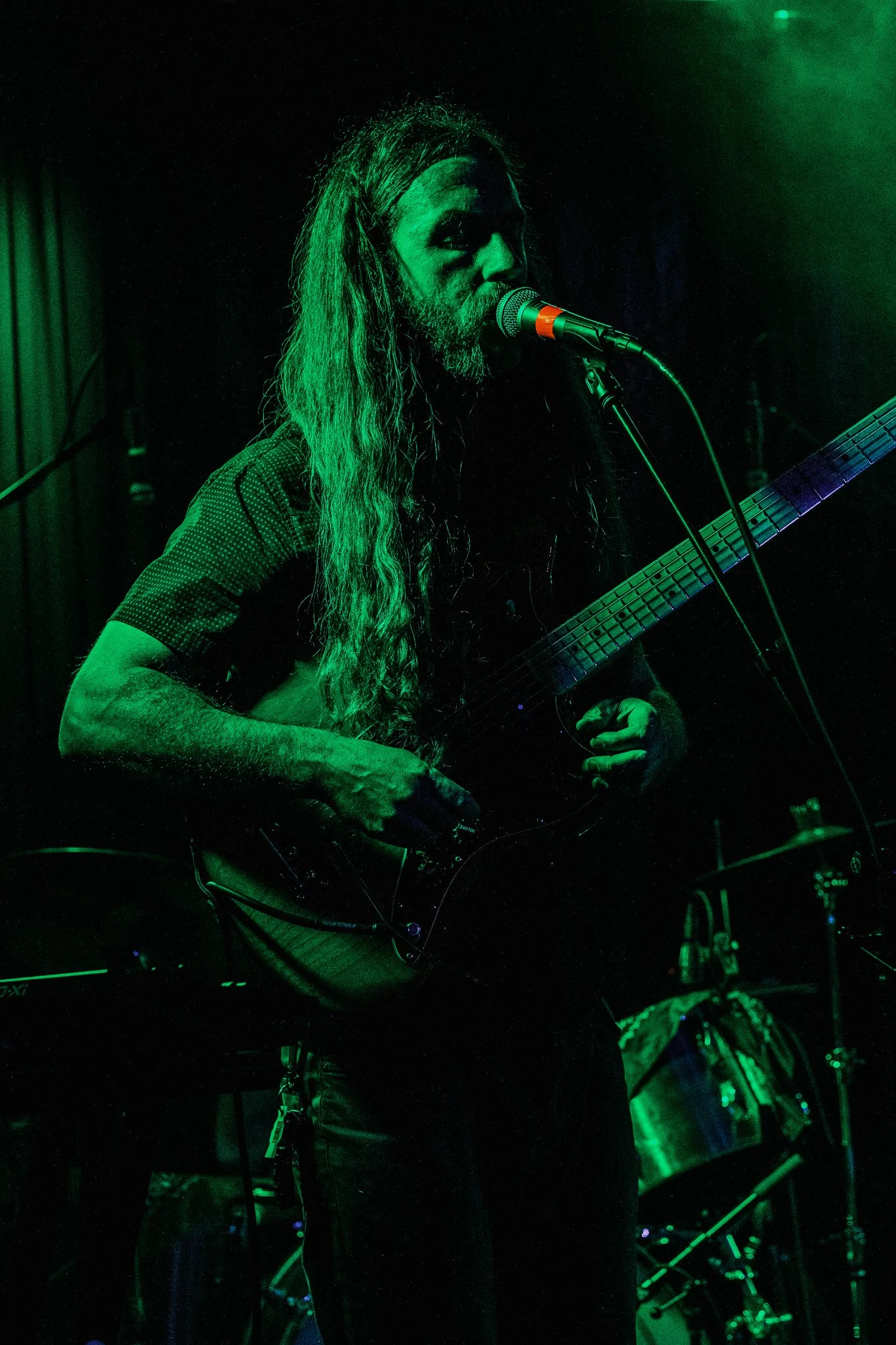 A man with long hair and a beard playing an electric guitar and singing into a microphone on stage, illuminated by green stage lighting, with a drum set in the background.