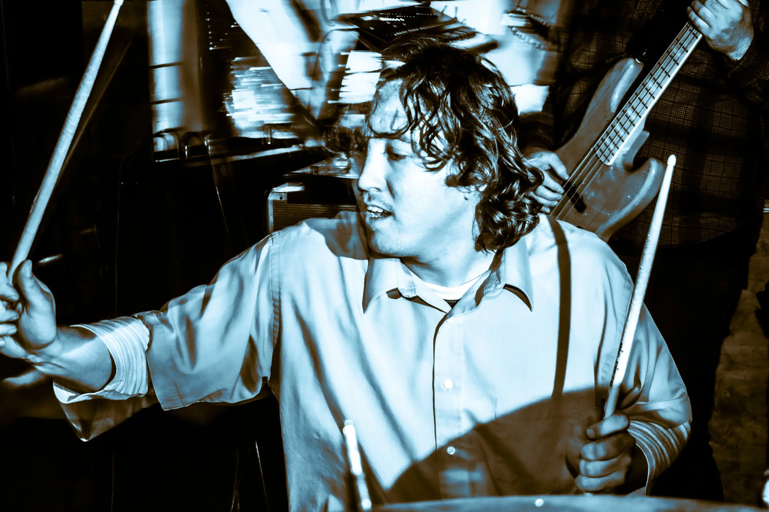 A young man with curly hair holding drumsticks, playing drums, and a person in the background playing a bass guitar, in a musical performance setting.