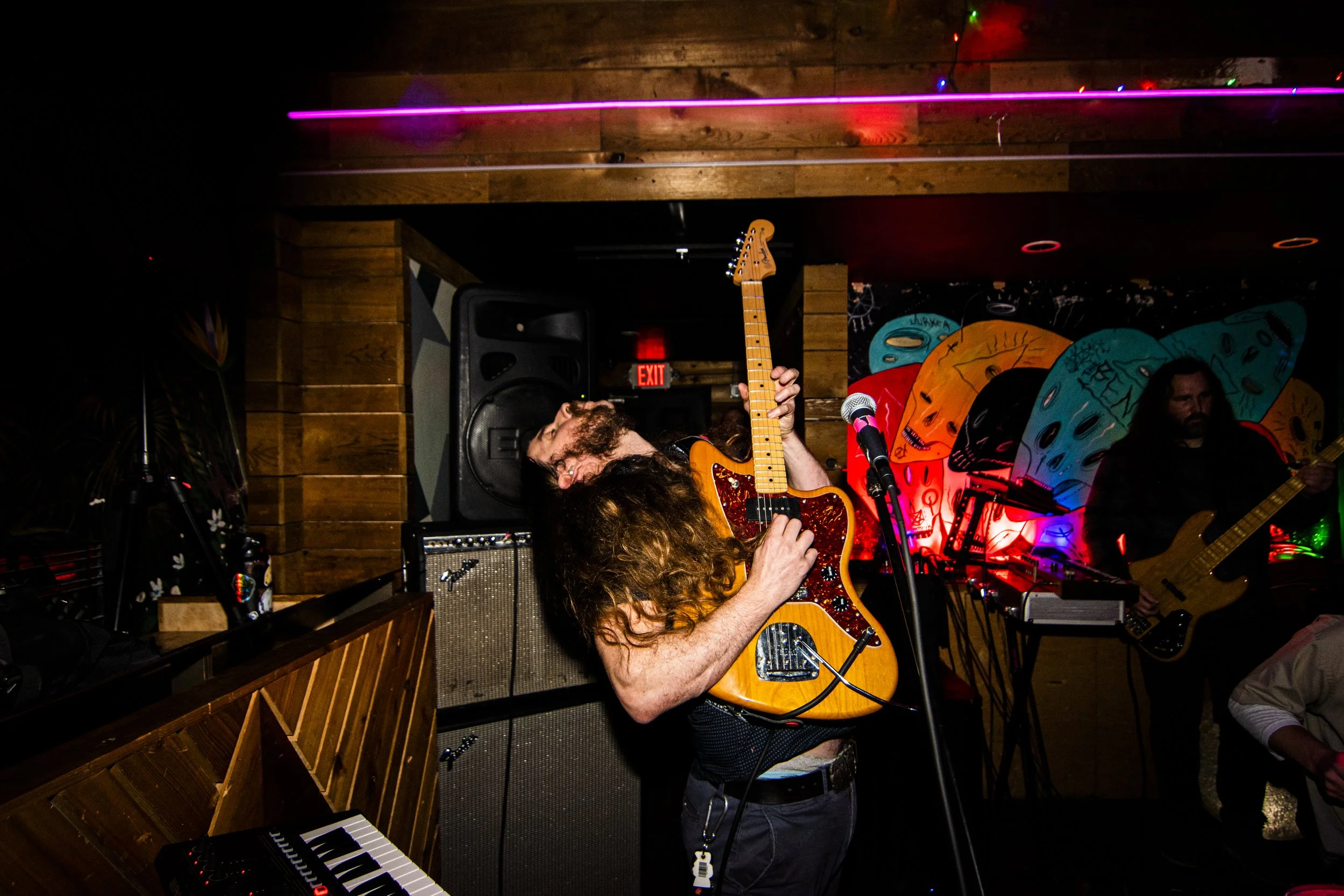 A musician with long hair and a beard, playing an orange electric guitar on stage at a live music venue with colorful butterfly artwork in the background.