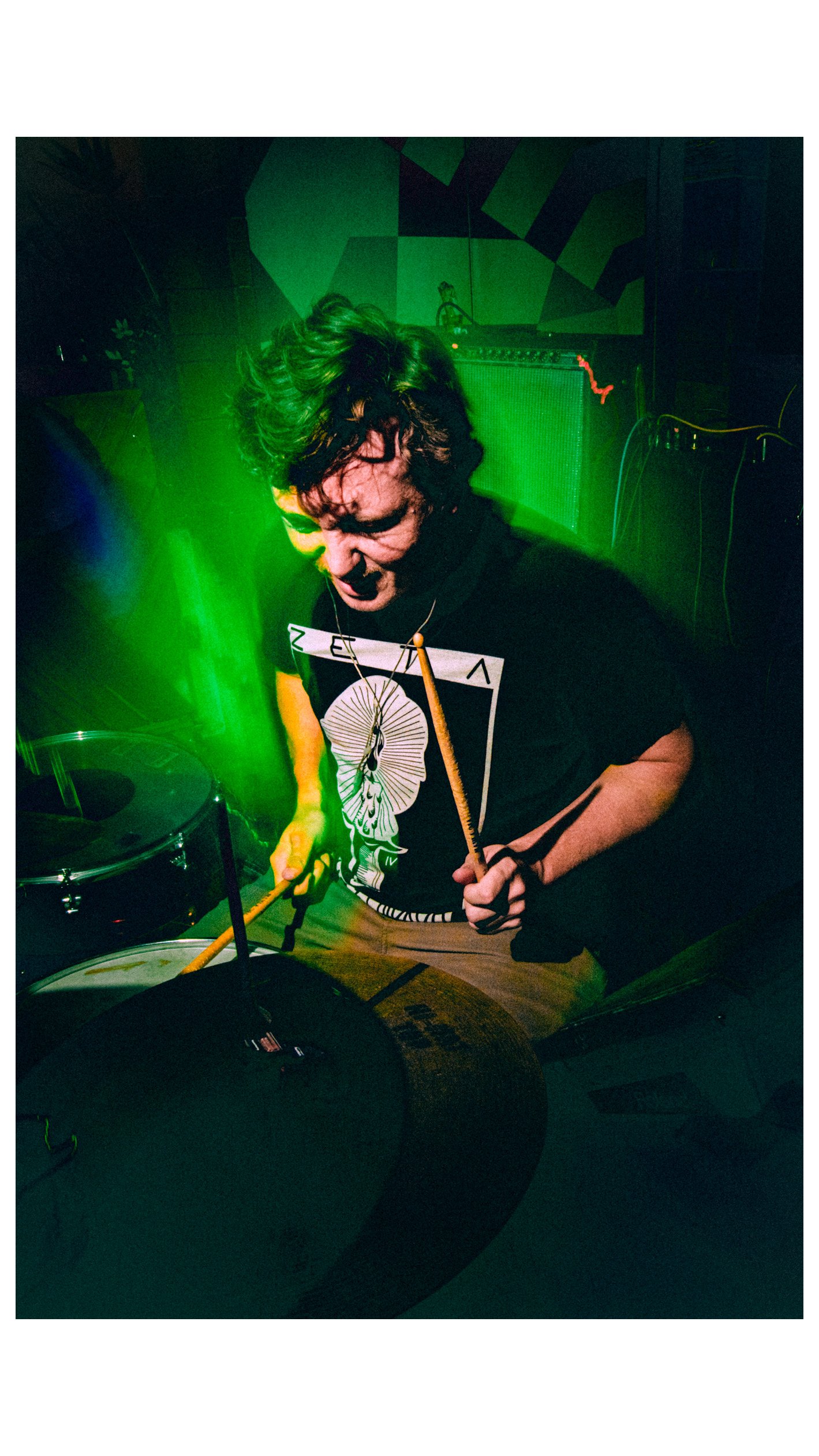 Rock band Liquid Pennies drummer Pierson Whicker playing drums in a dimly lit room with green lighting, wearing a graphic t-shirt featuring a butterfly and geometric patterns.