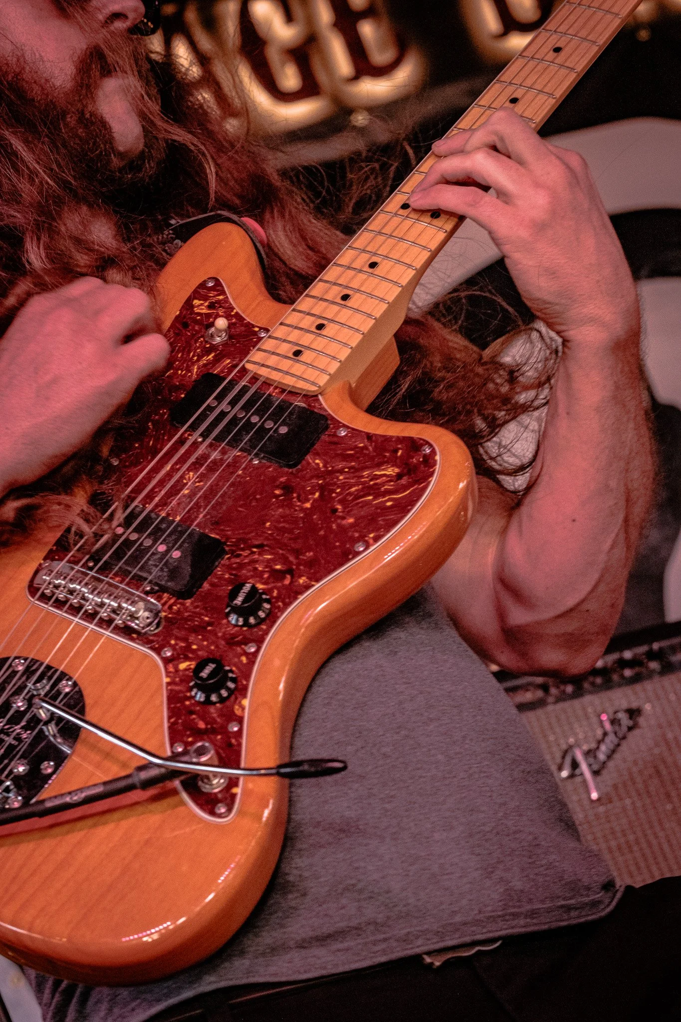 Close-up of Liquid Pennies guitarist Chas Binns with long hair playing a Fender Jazzmaster guitar with a tortoise shell pickguard, wooden body, and yellow neck at a music venue.