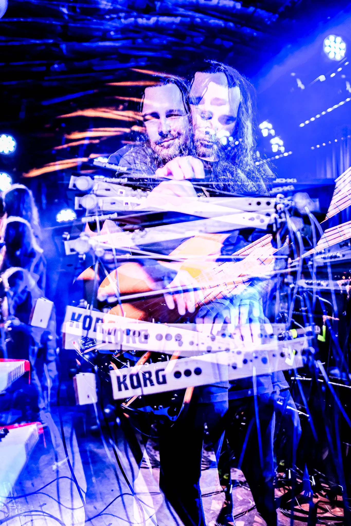 A double exposure photograph of two musicians on stage, one playing an electric guitar and the other reaching out with their hand, with stage lighting creating a vibrant, colorful glow.