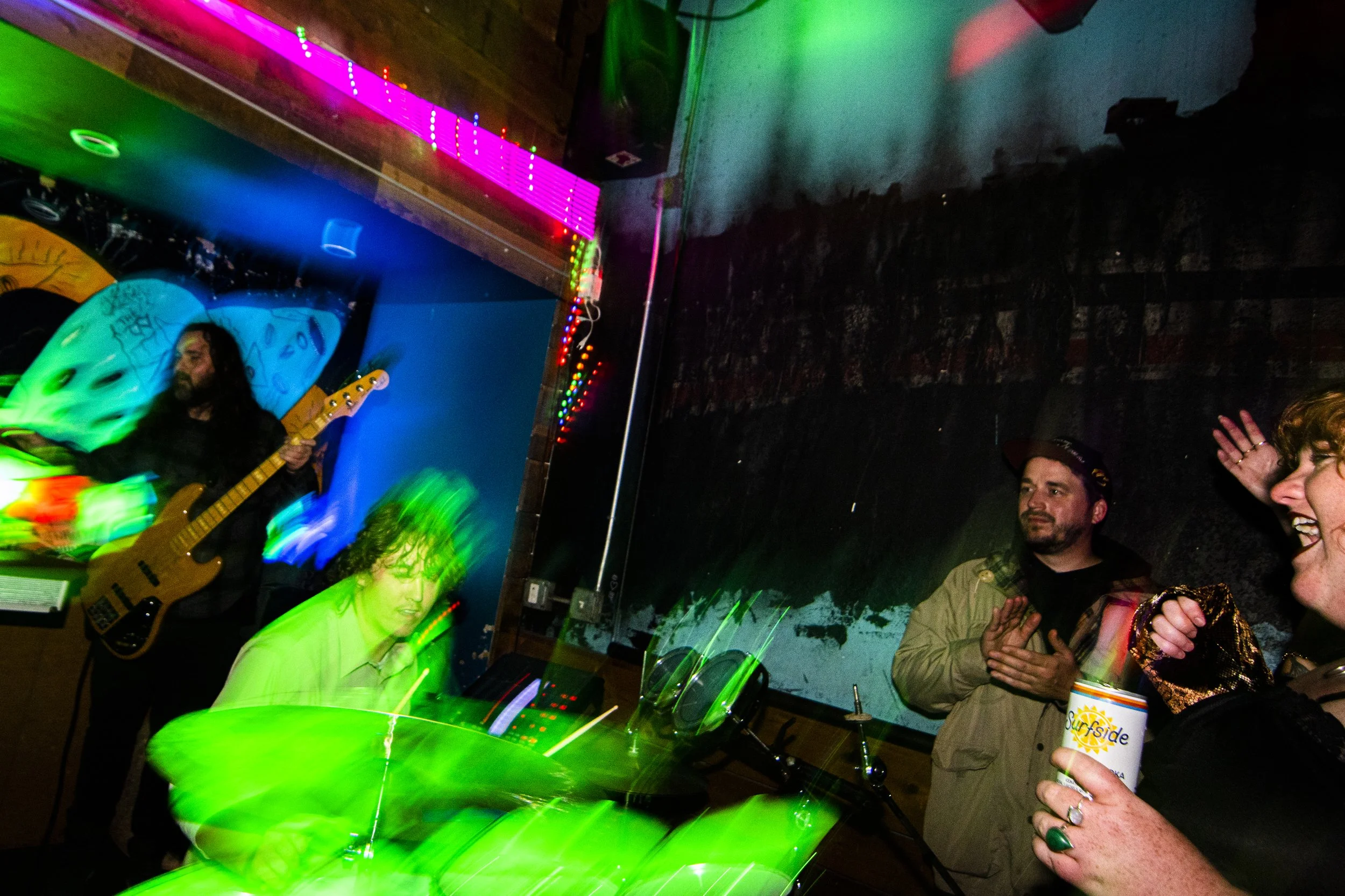 People enjoying a live music performance in a dimly lit venue with colorful lights, a guitarist playing on stage, and someone holding a drink while smiling.