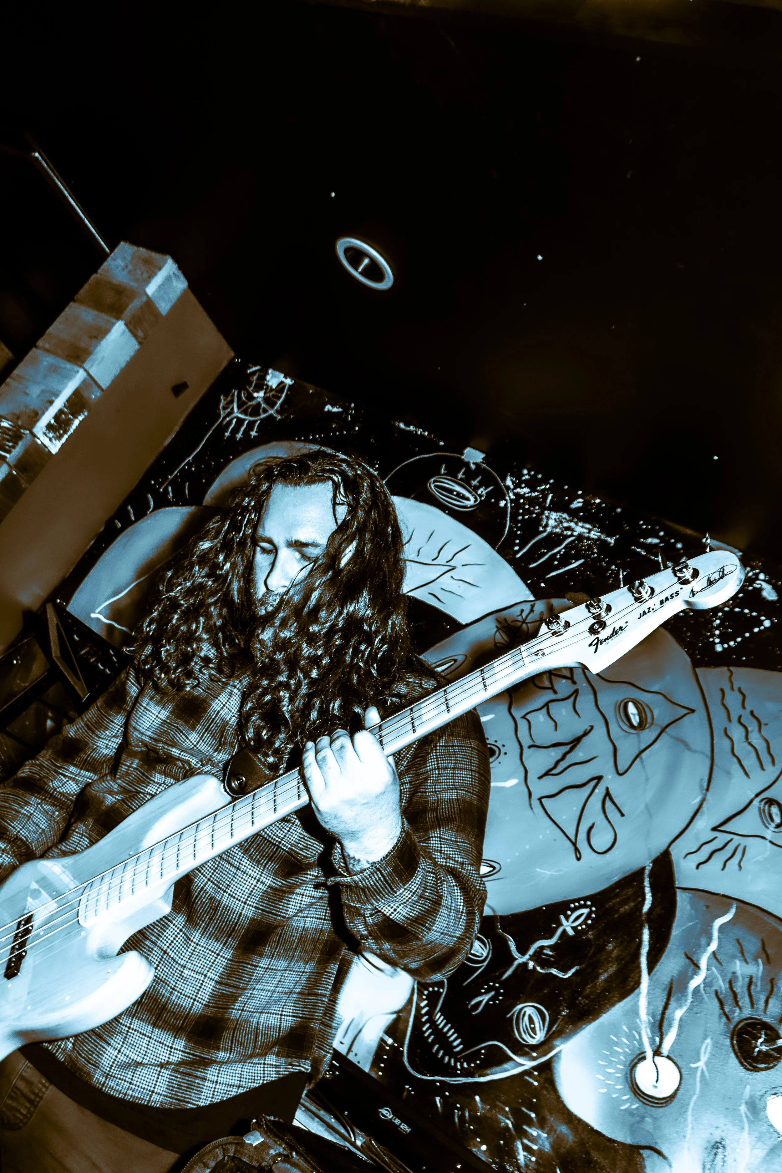 A person with long curly hair playing a bass guitar in front of a vibrant, artistic mural with celestial and abstract designs.