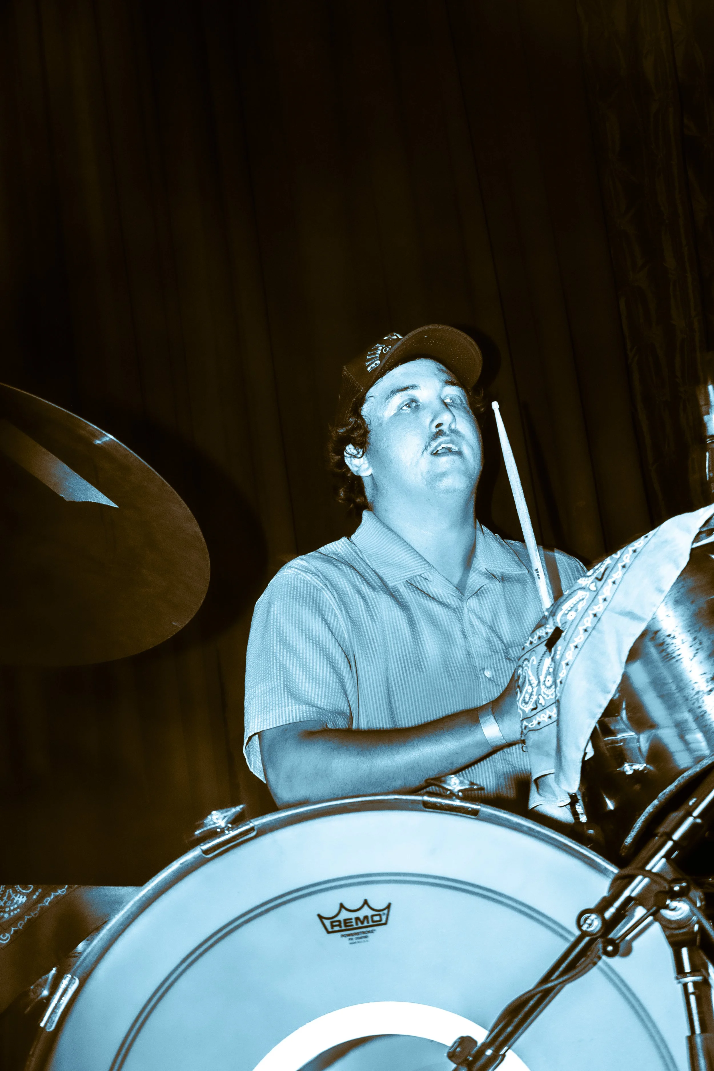 A man playing a drum set, holding a drumstick near his face, wearing a baseball cap and a striped shirt, with a dark curtain background.