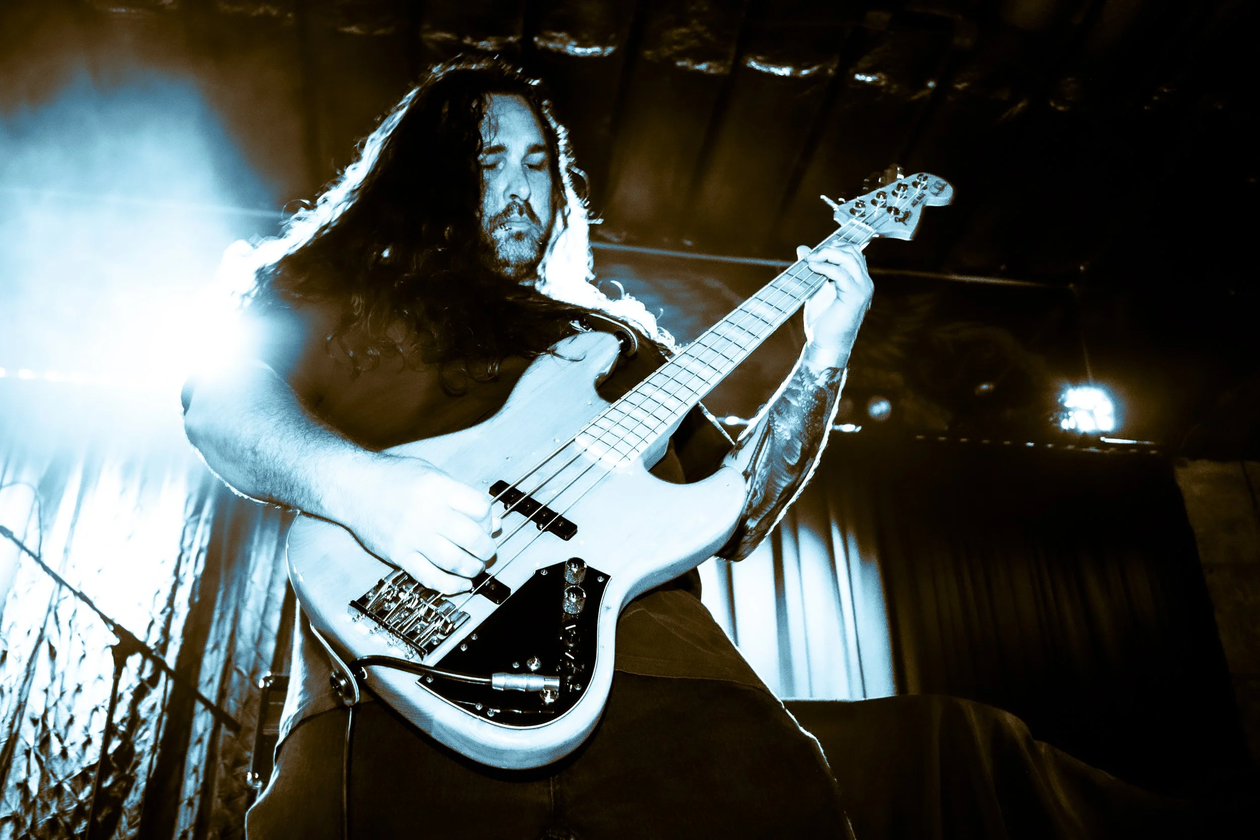 A man playing an electric bass guitar on stage with bright lights and dark curtains in the background.