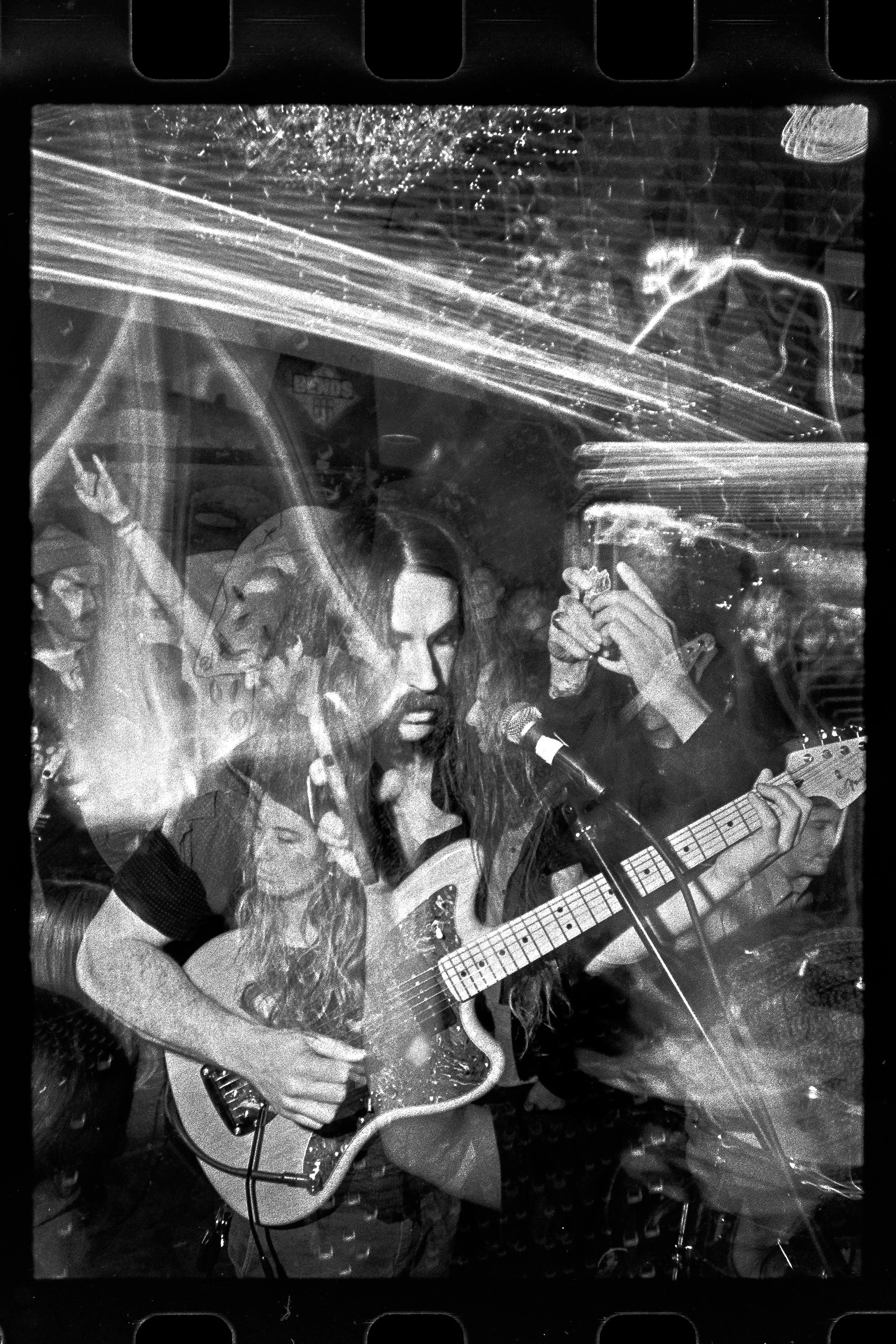 Black and white photo of a young woman playing electric guitar and singing into a microphone at a crowded music venue, with audience members and reflections visible.