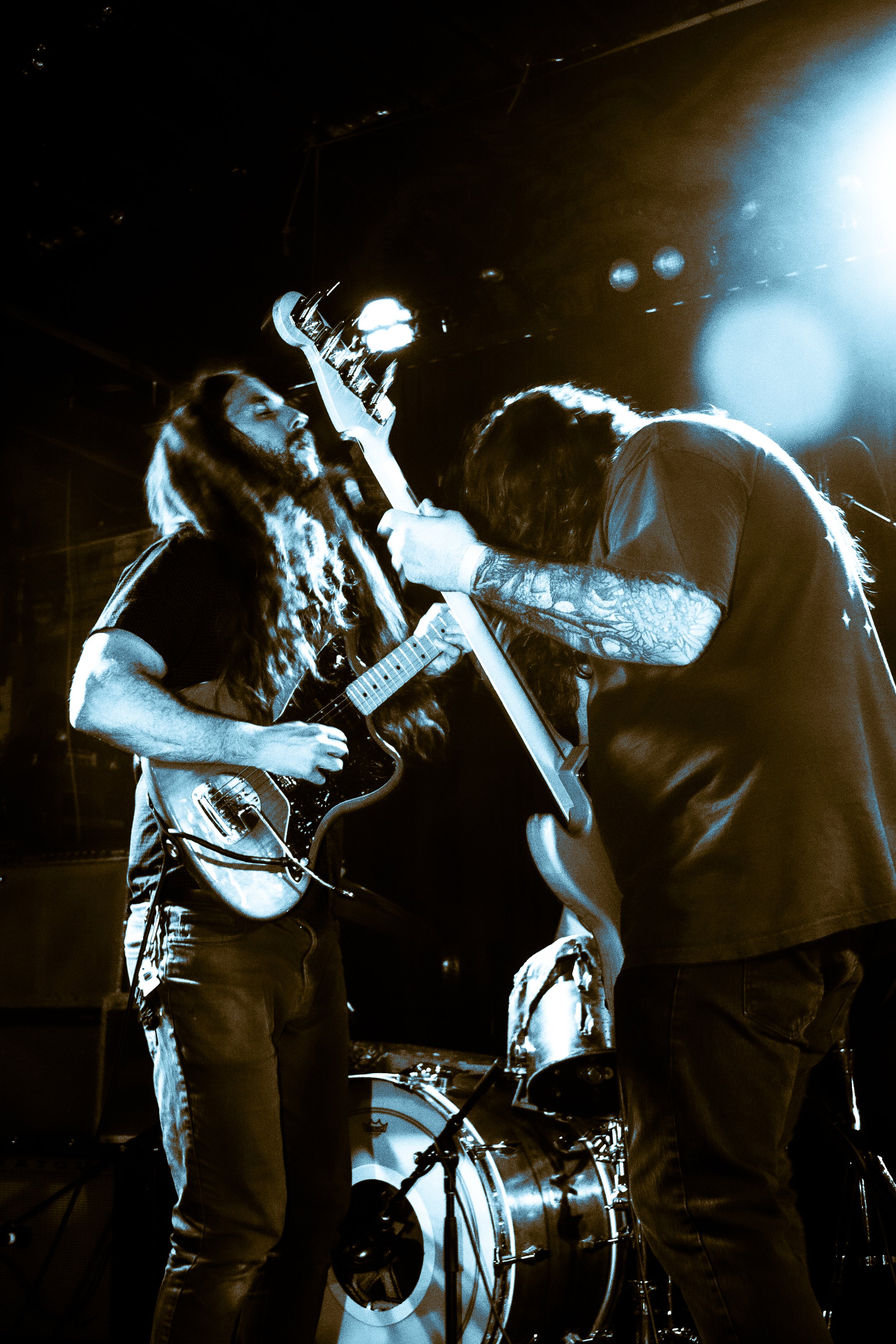 Two musicians performing on stage, both playing guitars. One has long hair and a beard, the other is headbanging with curly hair. The scene is illuminated by stage lights.