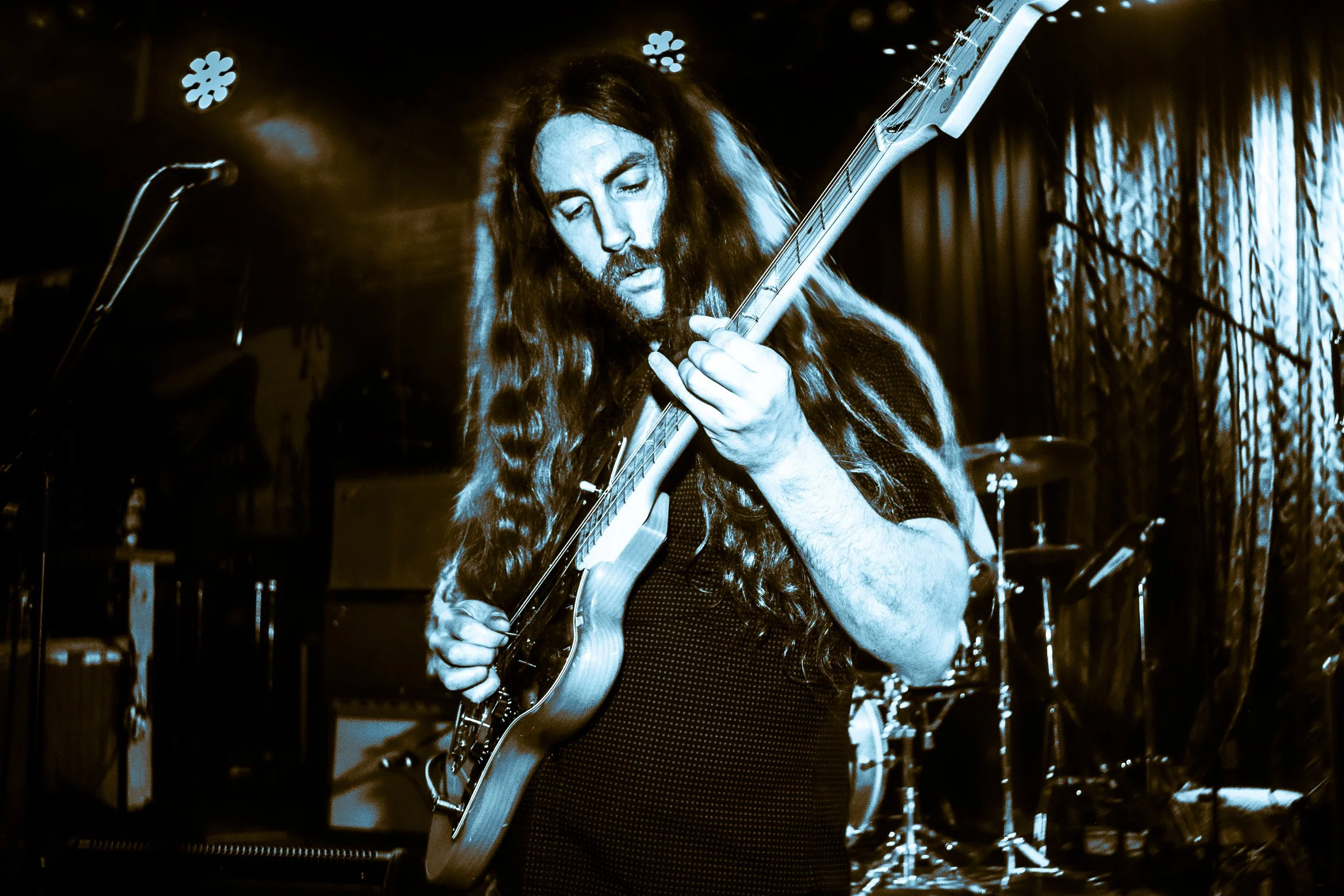 Rock band Liquid Pennies frontman/guitarist Chas BInns playing an electric guitar on stage, with drums and musical equipment in the background, illuminated by stage lights. From the LIquid Pennies official website