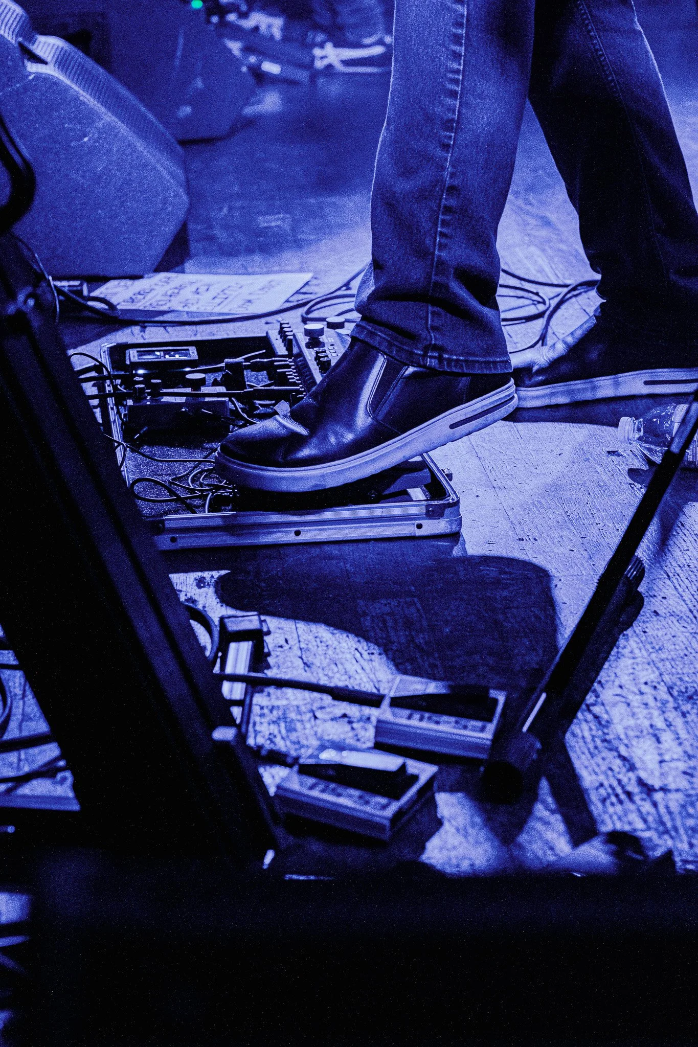 Person wearing black shoes and jeans standing on a pedalboard with guitar effects pedals, surrounded by cables on a wooden floor.