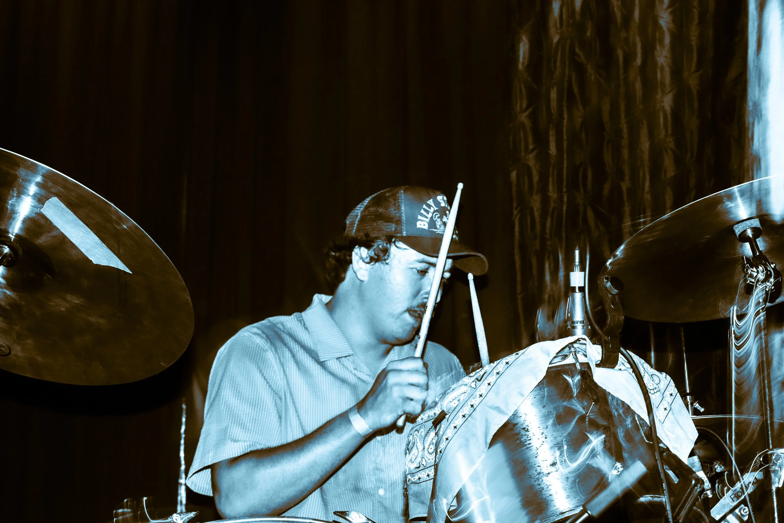 A person playing drums on stage, wearing a baseball cap and a short-sleeved shirt, surrounded by drum cymbals and equipment, with a dark background and curtain.