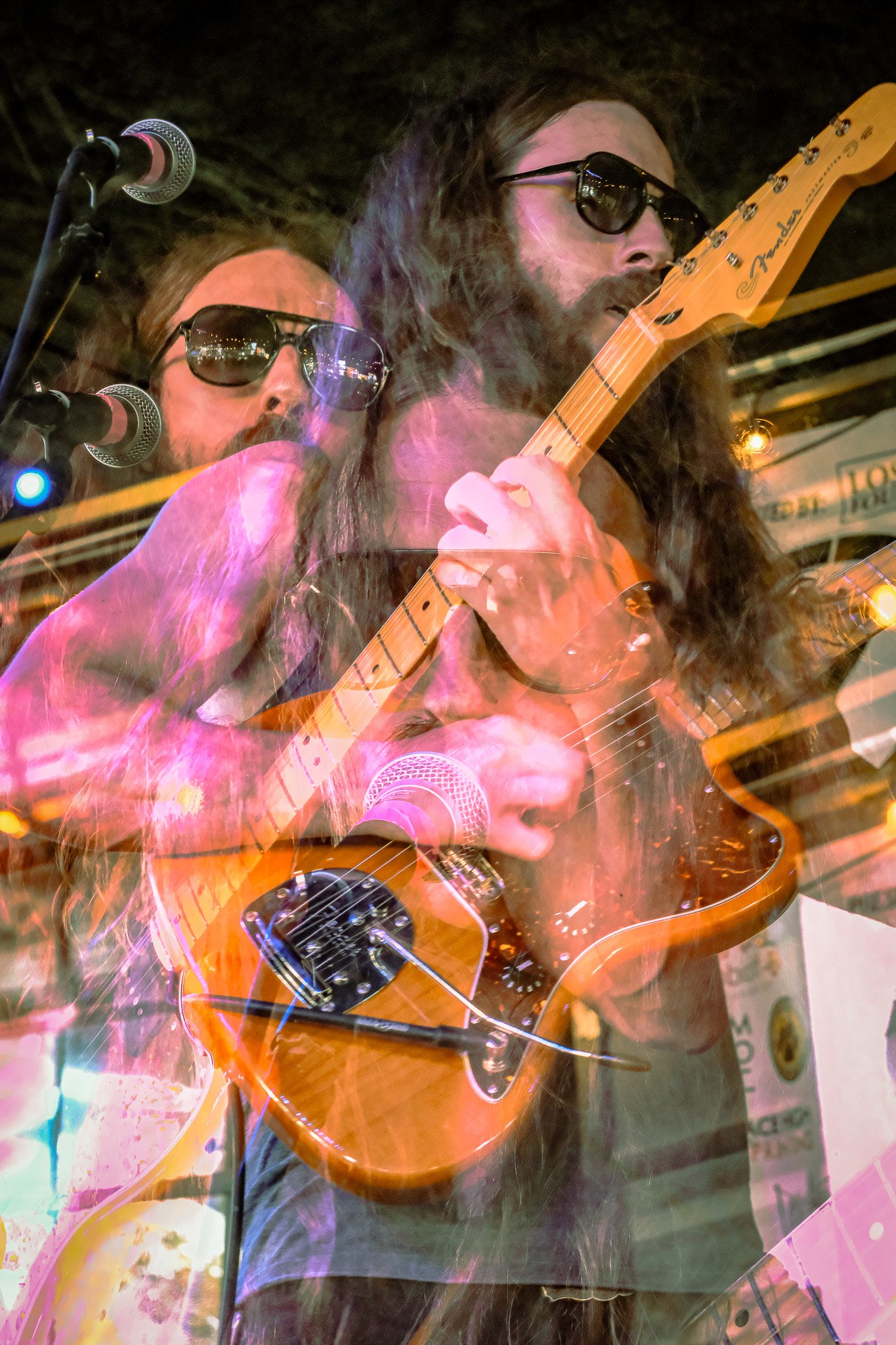 A person with long hair and sunglasses playing an acoustic guitar on stage. Multiple images of the person are superimposed, creating a layered effect.