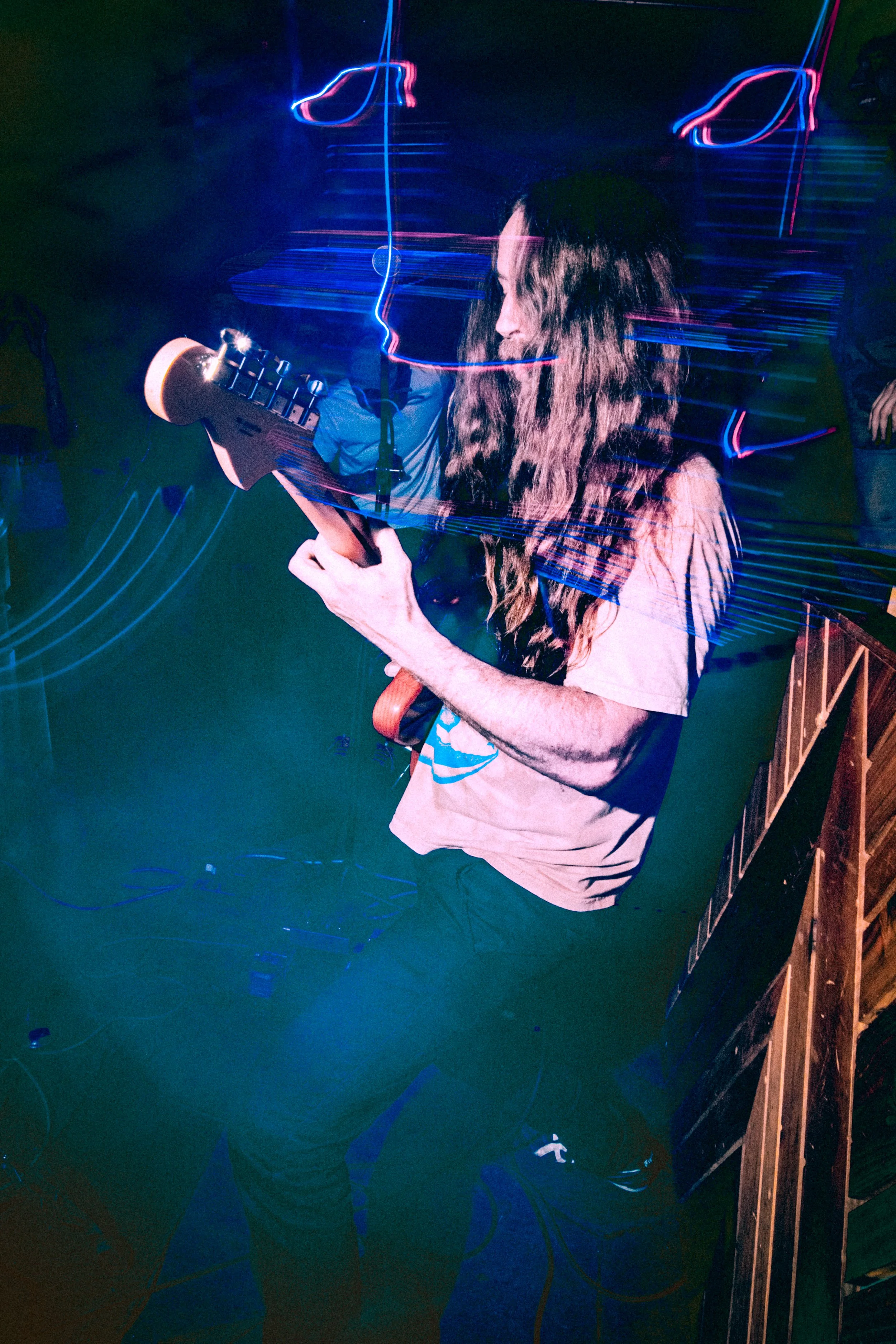 A man with long hair playing an electric guitar in a dimly lit room with blue and red light streaks.