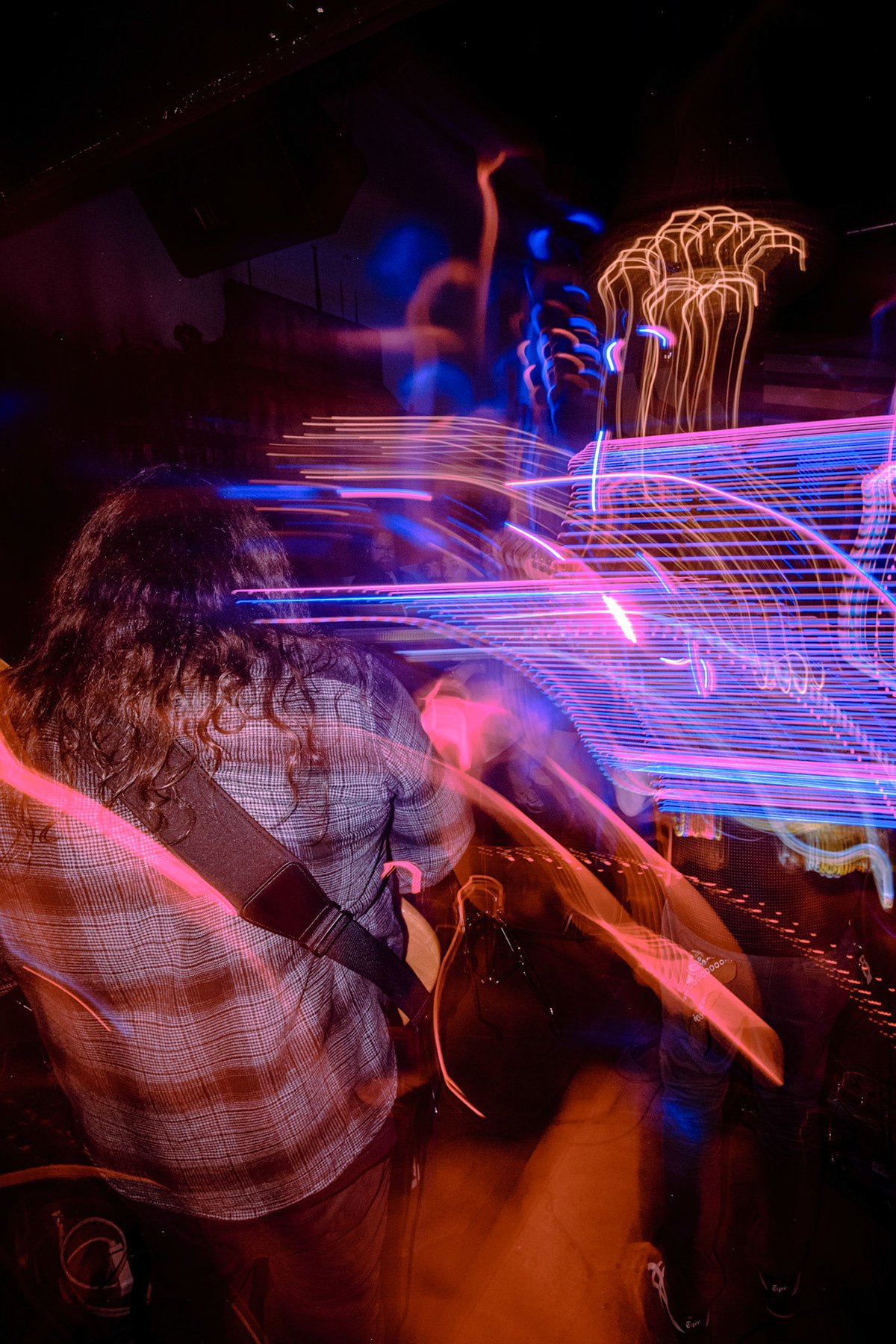 A person with long curly hair wearing a plaid shirt with a guitar strap stands in a dark room with colorful light trails creating a neon-like effect.