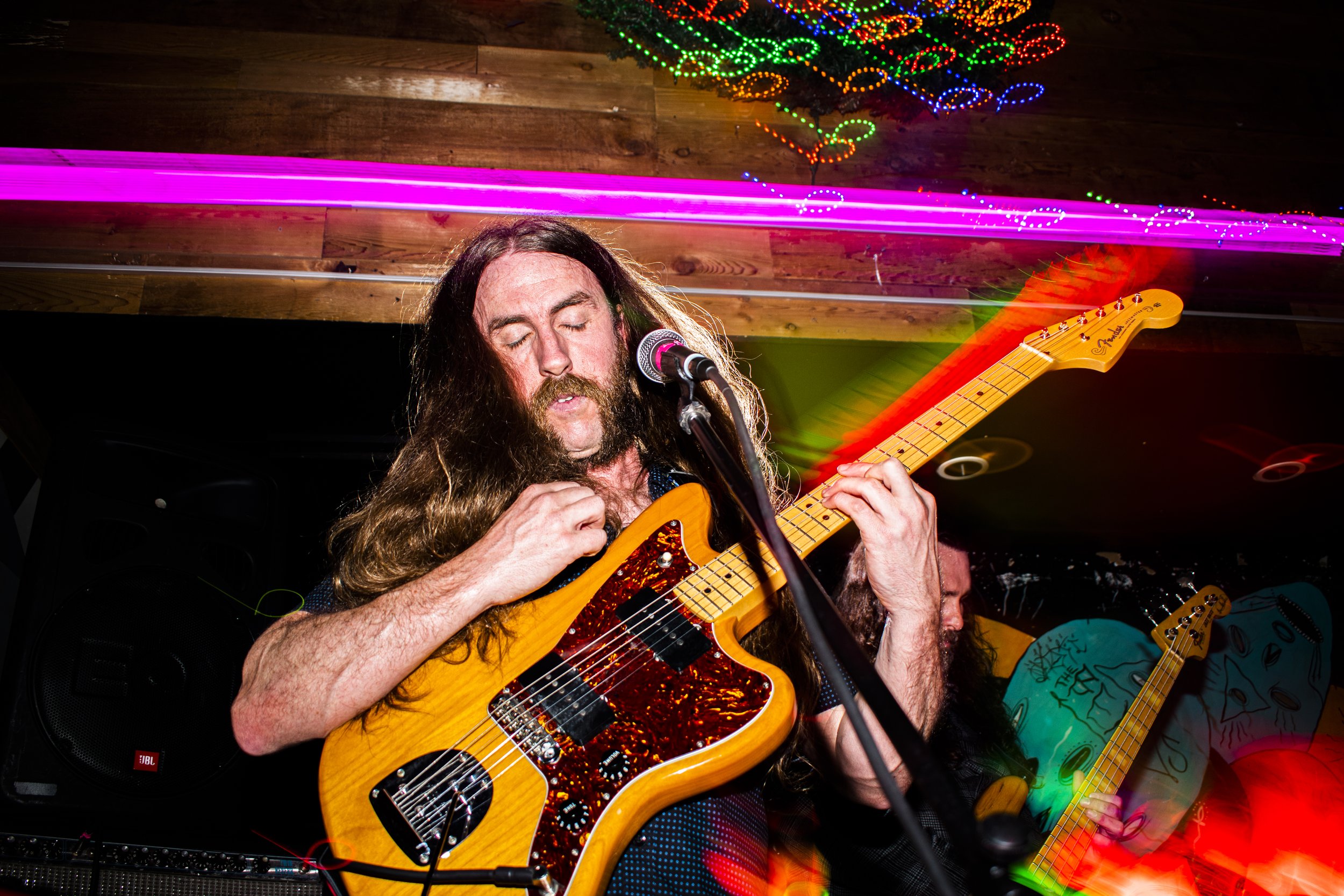 Liquid Pennies frontman/guitarist Chas Binns playing an electric guitar and singing into a microphone on stage with colorful lights and a wooden ceiling in the background.