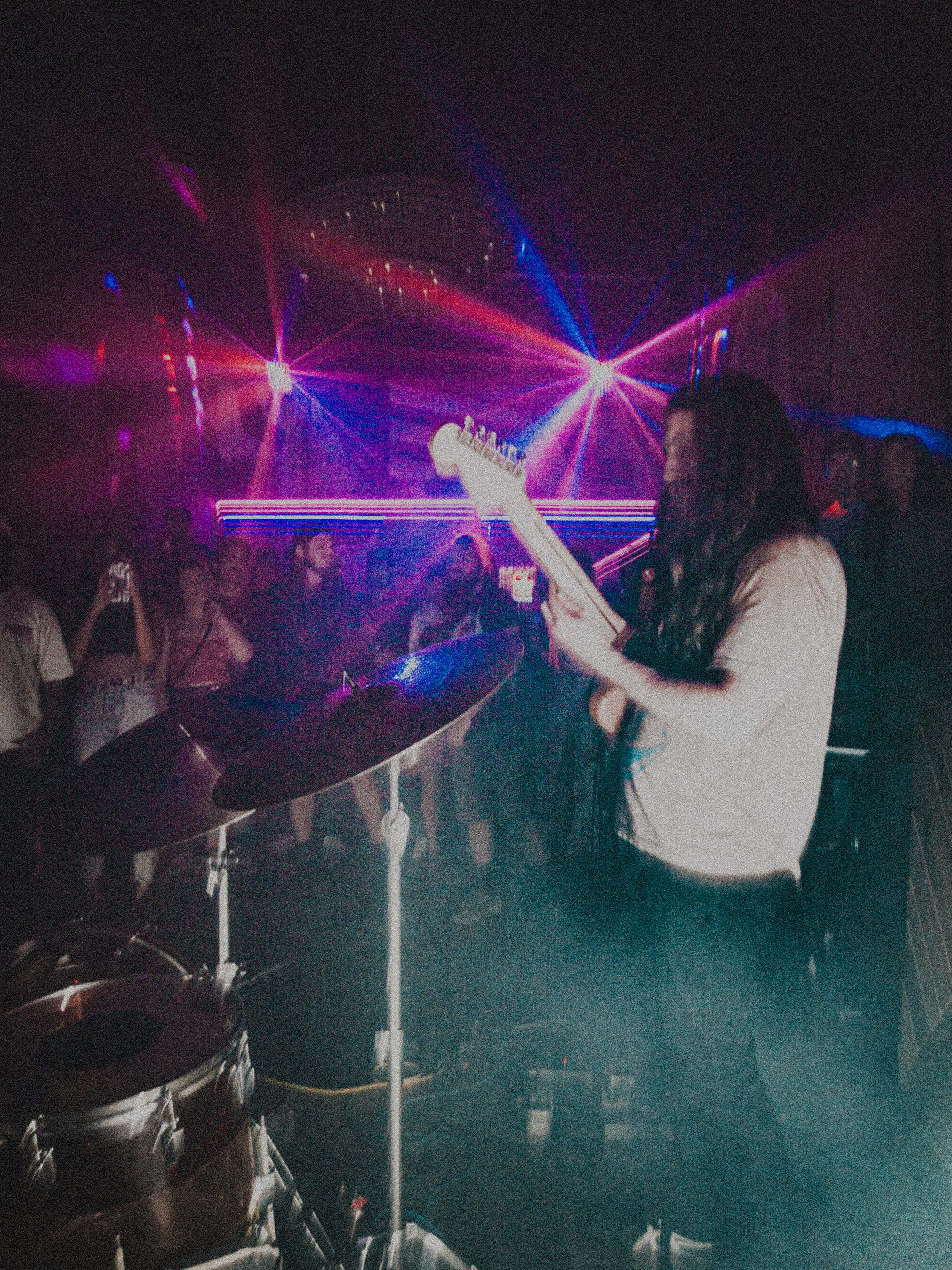 A woman playing the electric guitar on stage in a club with colorful laser lights, and a drum set nearby, while a crowd watches.