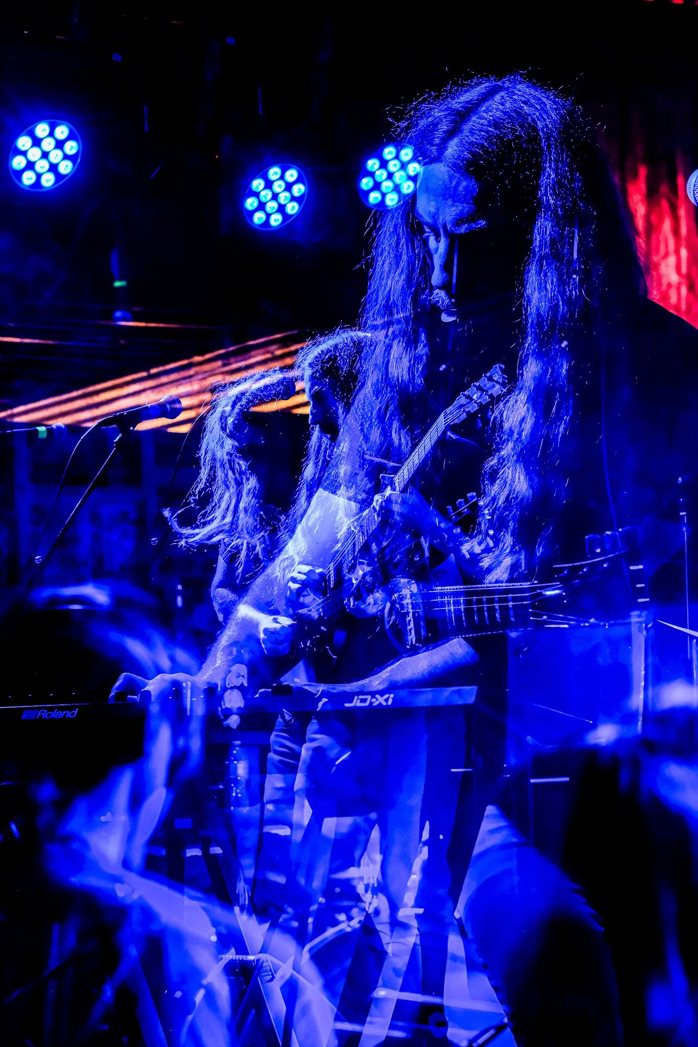 Musician playing guitar and keyboard on stage under blue and purple lights with long hair, with multiple exposure effect creating a layered appearance.
