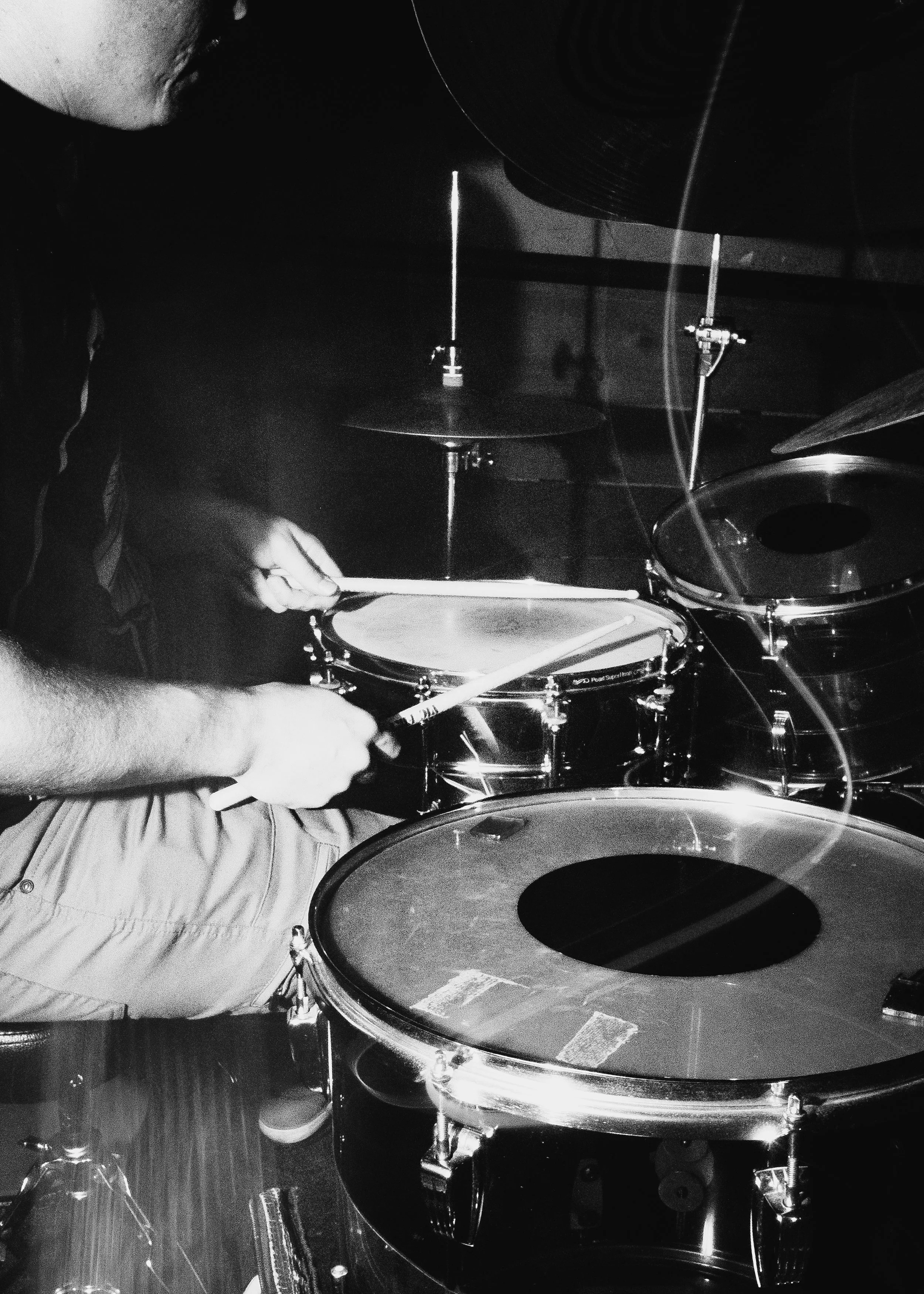 Black and white photo of a person playing the drums, hands gripping drumsticks, surrounded by drum equipment.