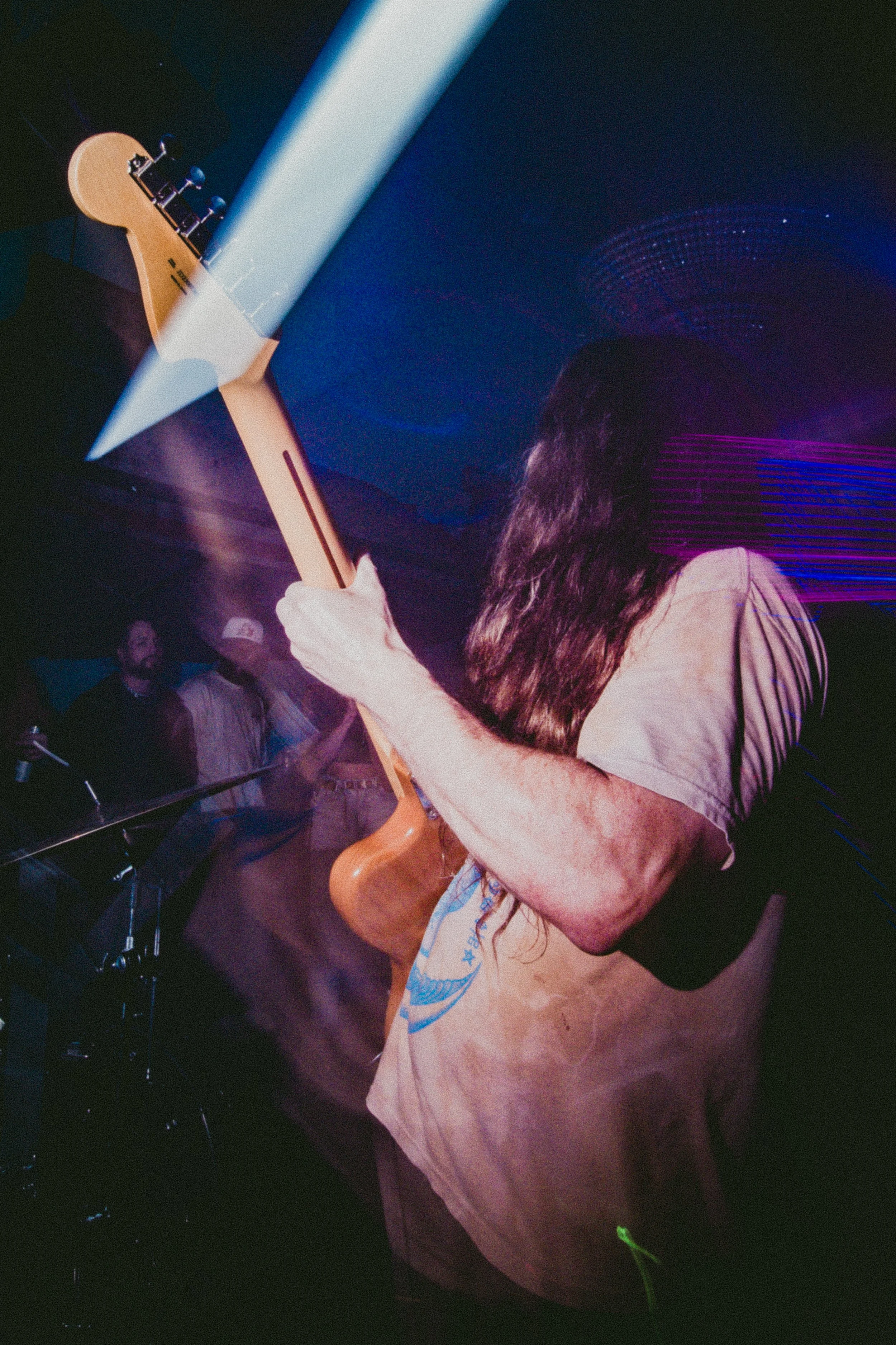 Person with long hair playing an electric guitar on stage with colorful lights and audience in the background.
