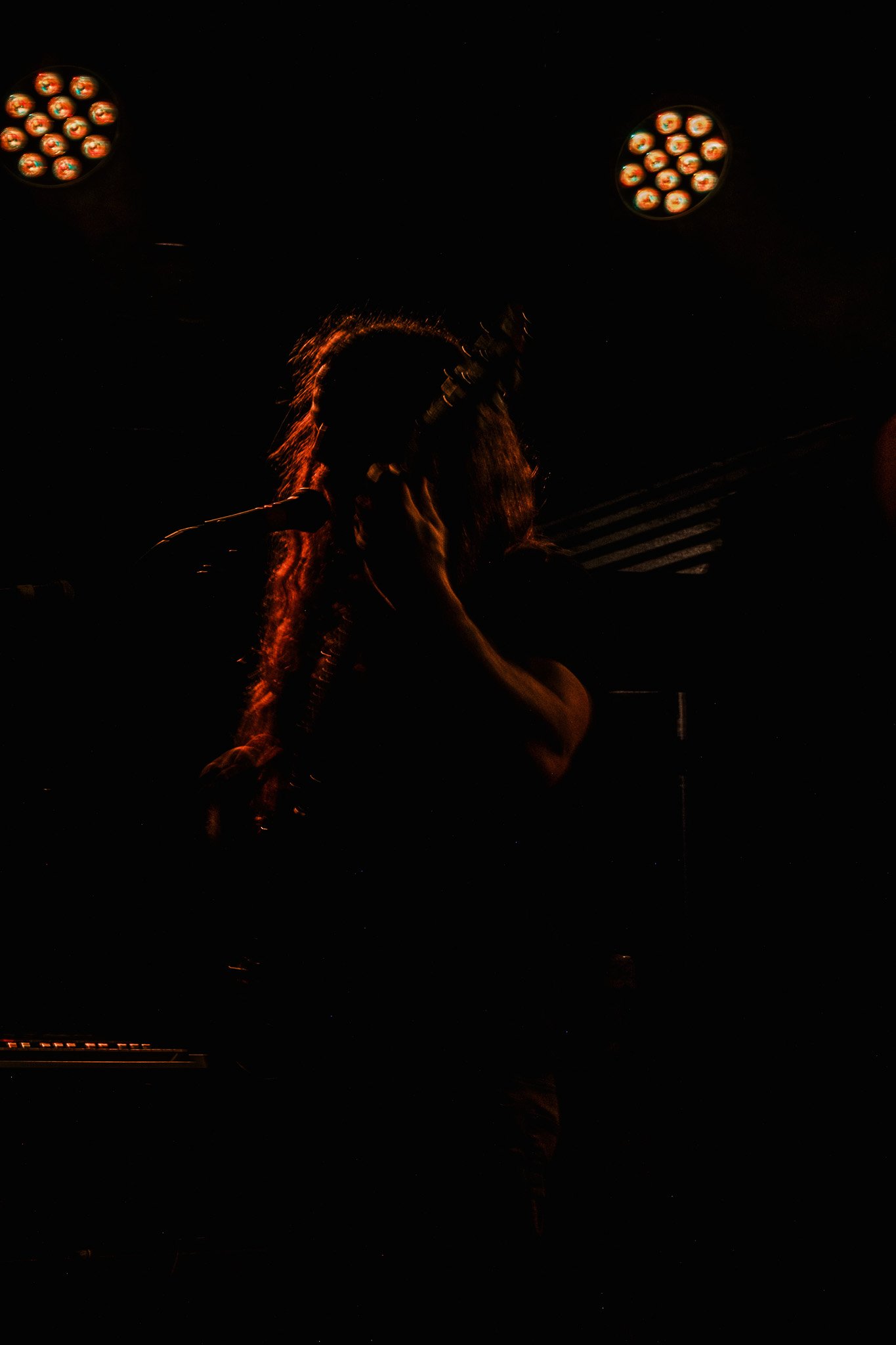 Dark silhouette of a person with long hair, holding a microphone, under stage lights.